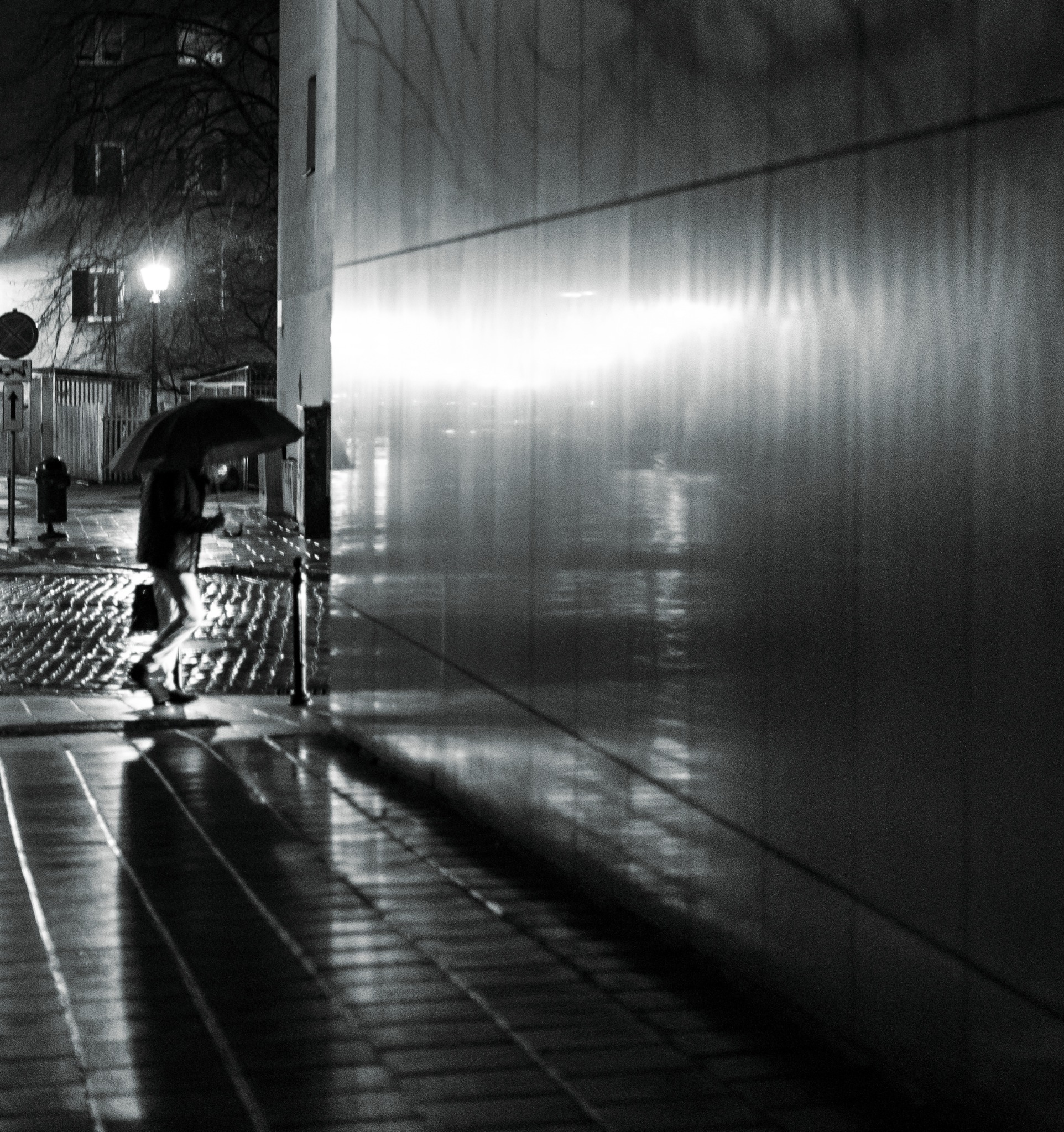 Czarno-białe zdjęcie nocne przedstawiające osobę z parasolem idącą chodnikiem wzdłuż ściany budynku odbijającej światła uliczne.