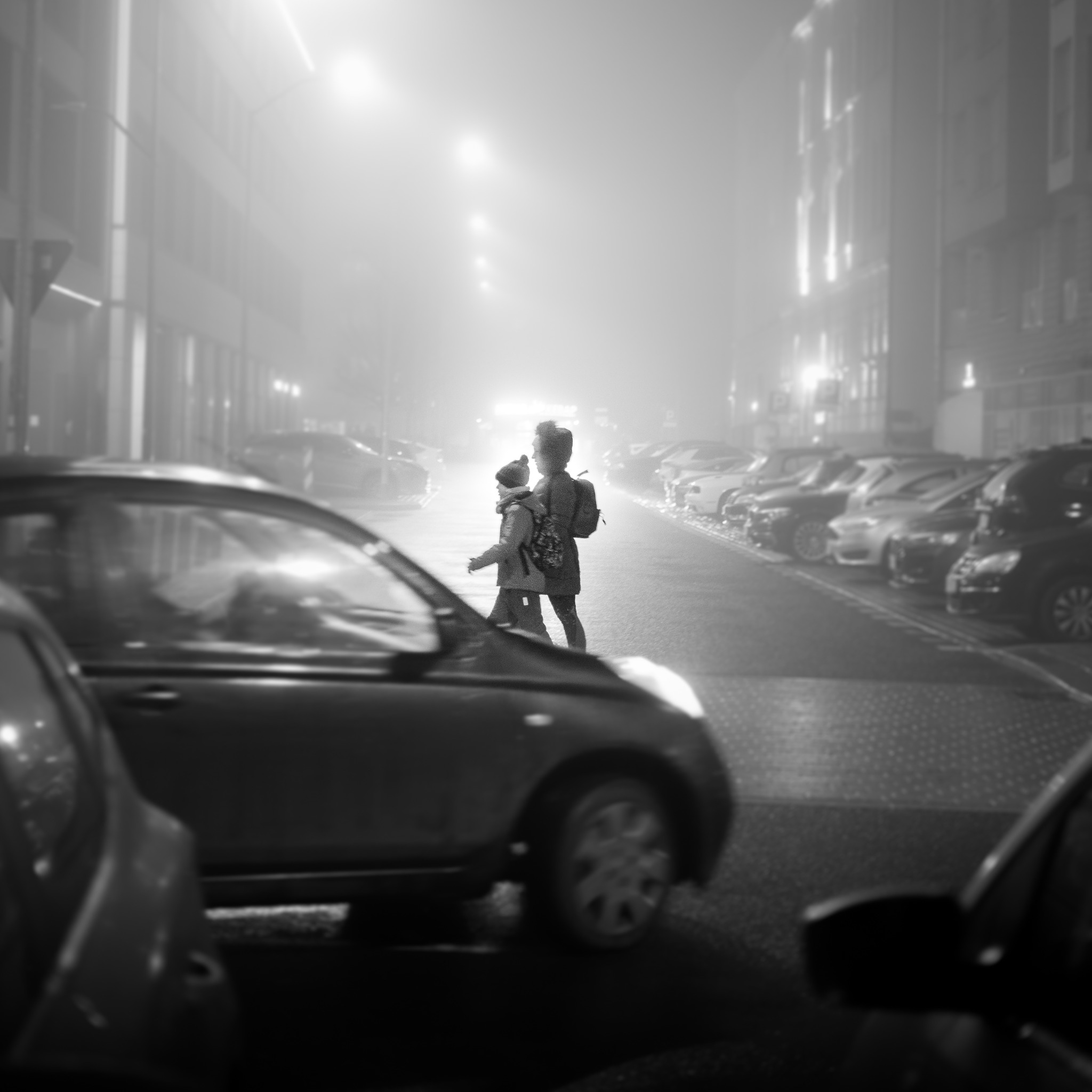 Czarno-białe zdjęcie przedstawia matkę z dzieckiem przechodzących przez ulicę w gęstej mgle, z rzędami zaparkowanych samochodów po obu stronach i światłami ulicznymi rozproszonymi przez mgłę.