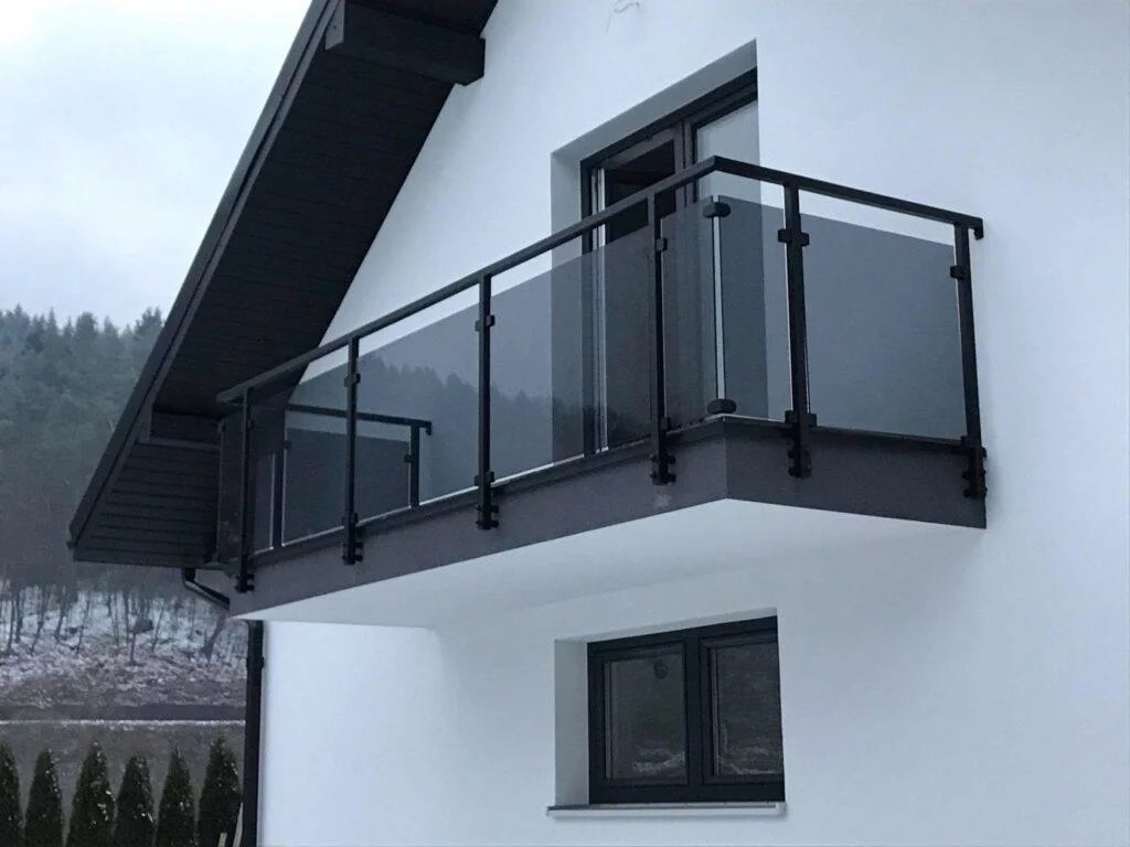 Nowoczesny balkon z czarną ramą i szklanymi panelami, widok z dołu na tle białej elewacji domu i zalesionego wzgórza w pochmurny dzień.