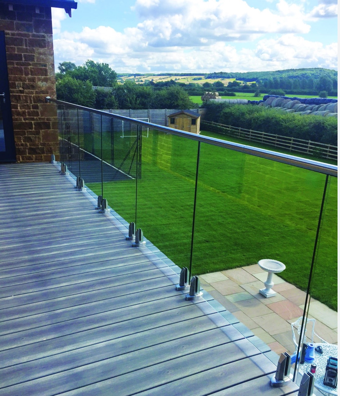 Szklana balustrada z mocowaniami punktowymi ze stali nierdzewnej na tarasie w Młodzieszynie, widok na zielony ogród z domkiem narzędziowym i polami w tle.