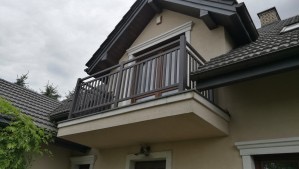 Balkon z ciemnobrązową, metalową balustradą o pionowych szczeblach, zamontowany na jasnobeżowej elewacji domu jednorodzinnego, widok z dołu na tle pochmurnego nieba i fragmentów zieleni.