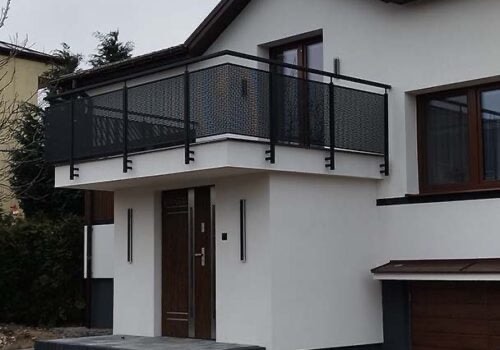 Czarna, metalowa balustrada balkonowa z ażurowym wypełnieniem na tle białej elewacji domu z brązowymi oknami i drzwiami wejściowymi.