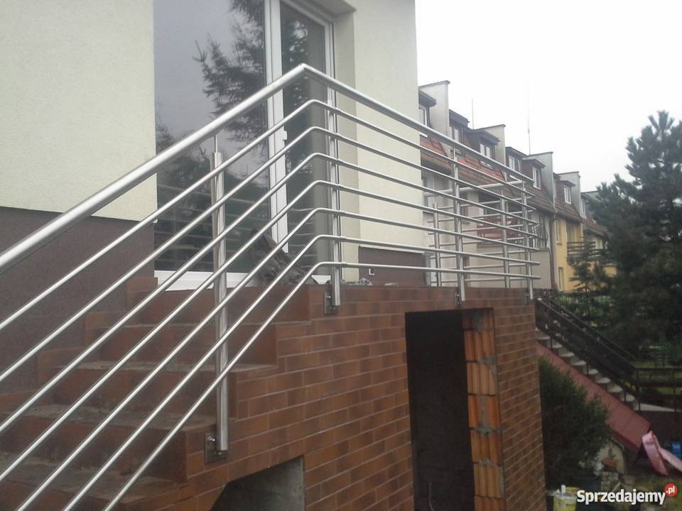 Zewnętrzne schody z cegły z prostą, poziomą balustradą ze stali nierdzewnej, widoczne budynki w tle.