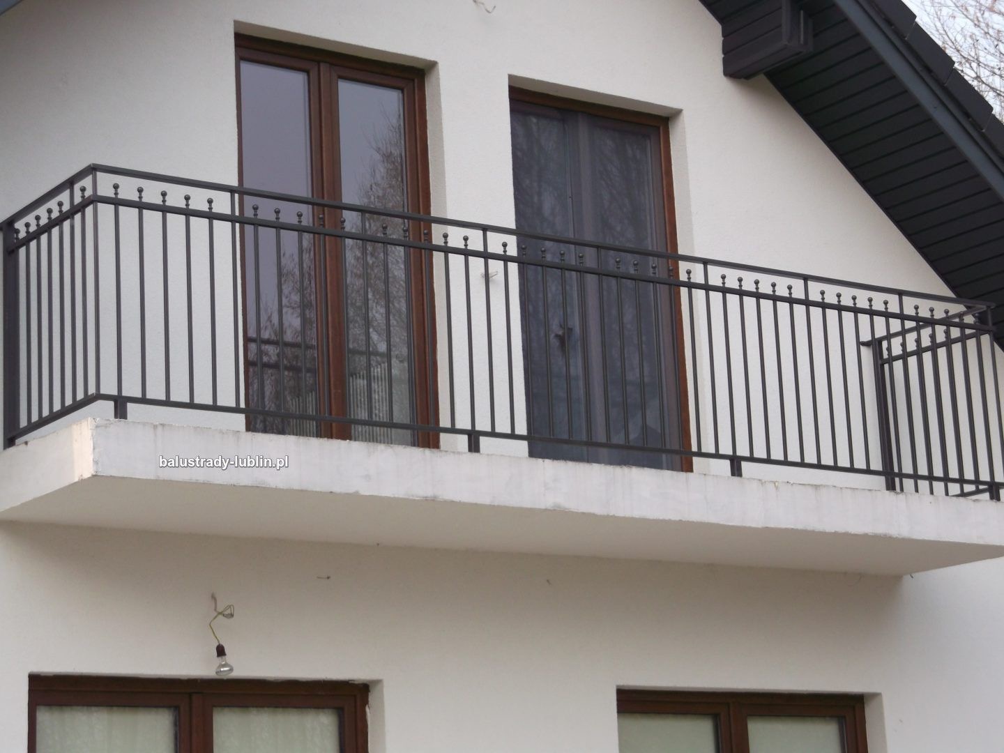 Czarna, metalowa balustrada balkonowa z pionowymi prętami i kulkami na górze, zamontowana na białym balkonie budynku z brązowymi oknami.