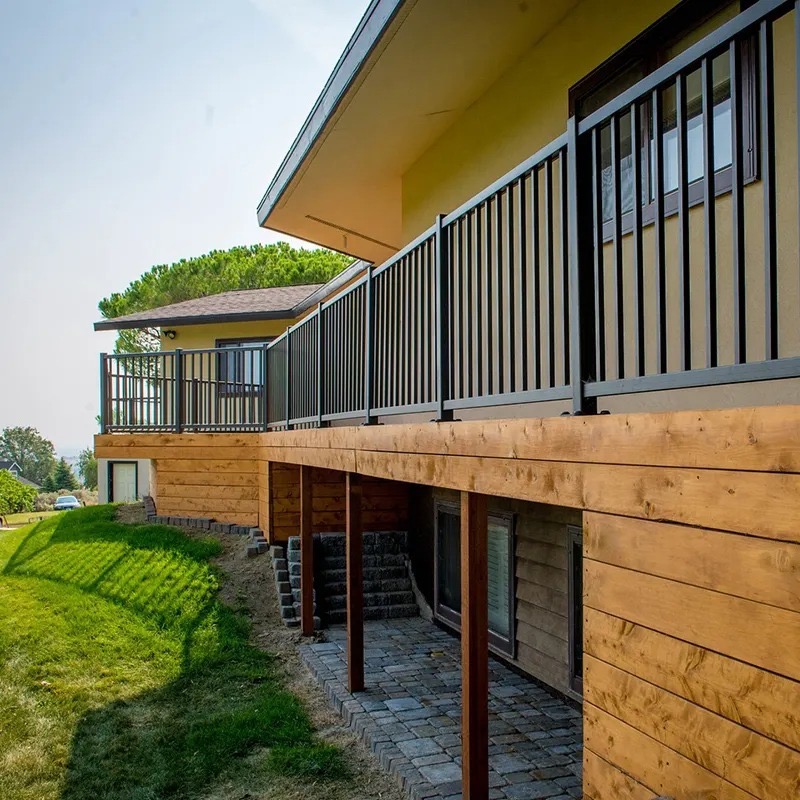 Nowoczesna, czarna balustrada pionowa na drewnianym tarasie domu z widokiem na zielony trawnik i oddalony krajobraz.