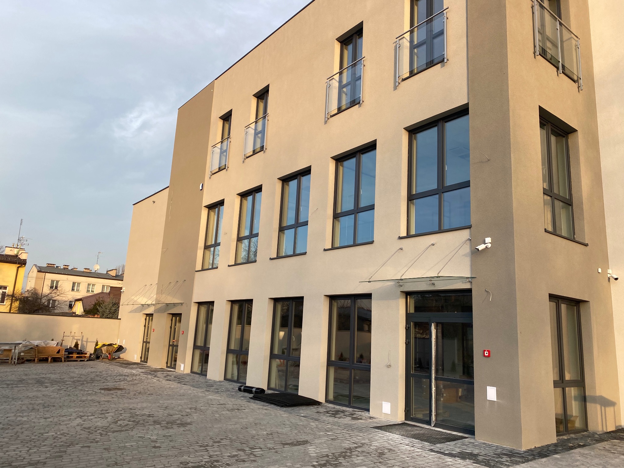 Nowoczesny budynek biurowy z beżową elewacją, czarnymi ramami okiennymi, szklanymi balustradami balkonowymi i przeszklonymi daszkami nad wejściami, widok z zewnątrz na brukowany plac.