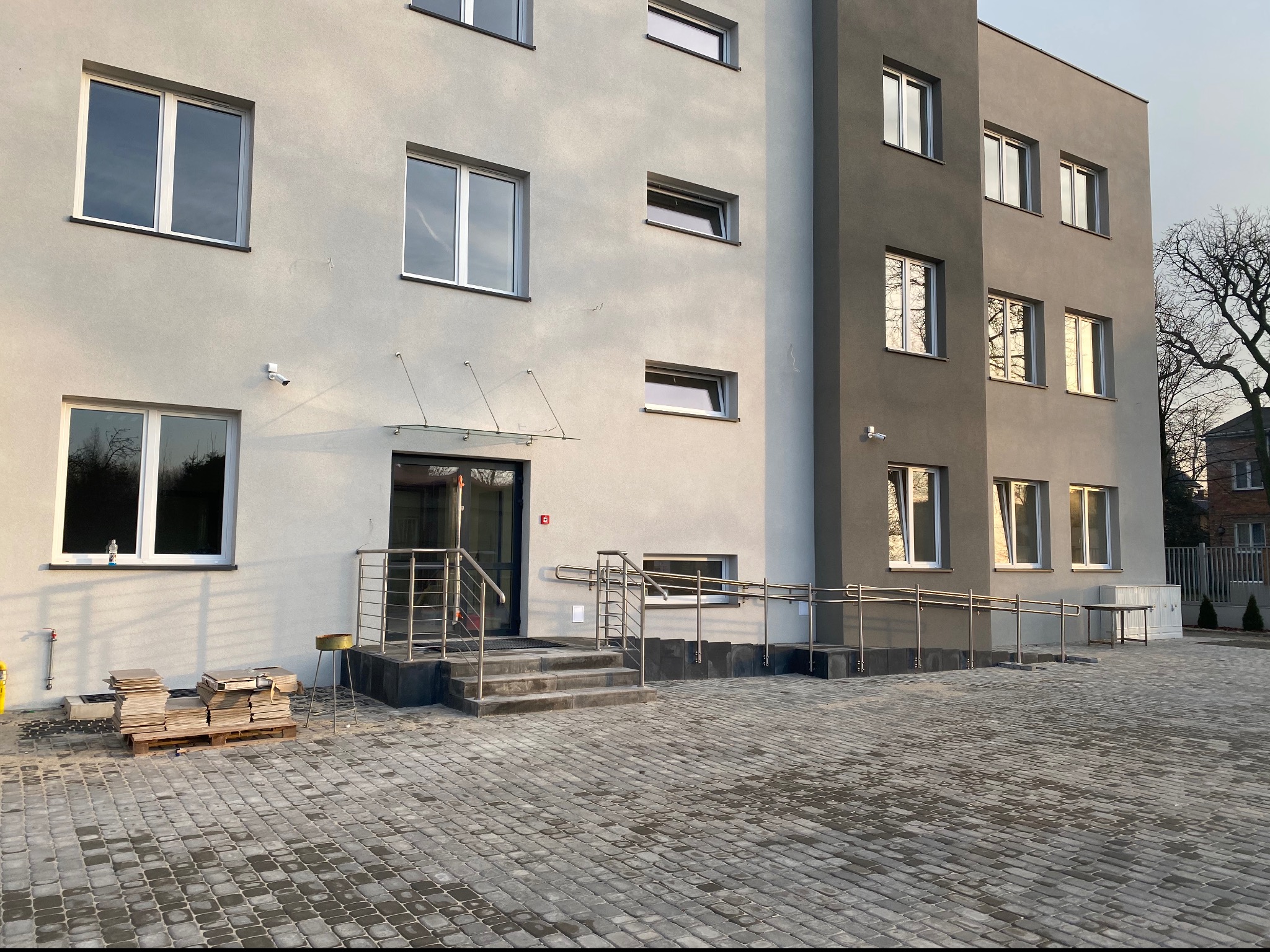 Nowoczesny budynek z jasną elewacją i szarą częścią boczną, z wejściem zabezpieczonym balustradą ze stali nierdzewnej i podjazdem dla osób niepełnosprawnych, na pierwszym planie ułożona kostka...
