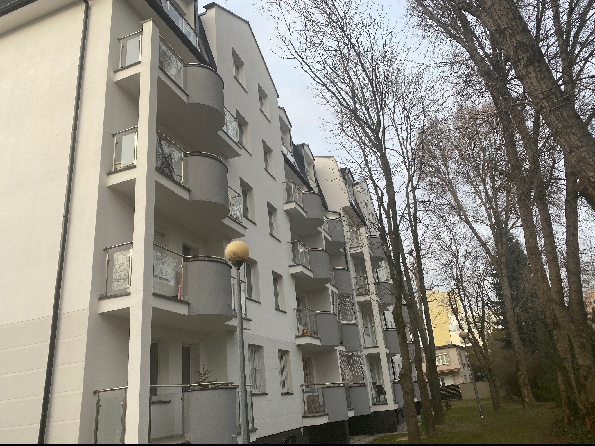 Nowoczesny budynek wielorodzinny z balkonami zabezpieczonymi szklanymi balustradami ze stalowymi elementami, widok z perspektywy trawnika z drzewami bezlistnymi.