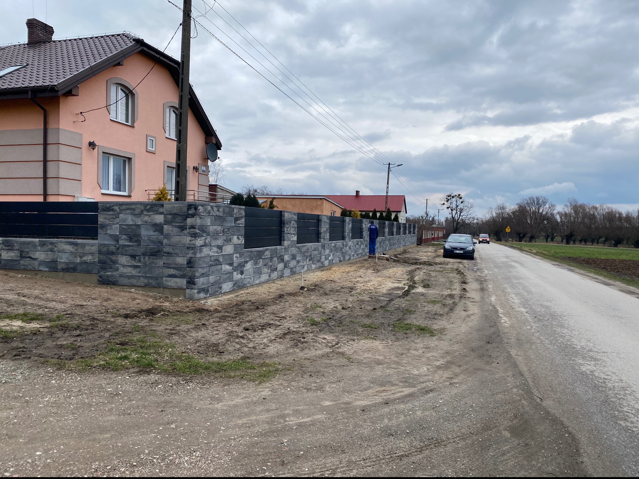 Nowoczesne ogrodzenie z szarej kostki łupanej z poziomymi czarnymi przęsłami, widoczne z perspektywy ulicy, z pracownikiem w niebieskim kombinezonie w tle i samochodem osobowym zaparkowanym obok.