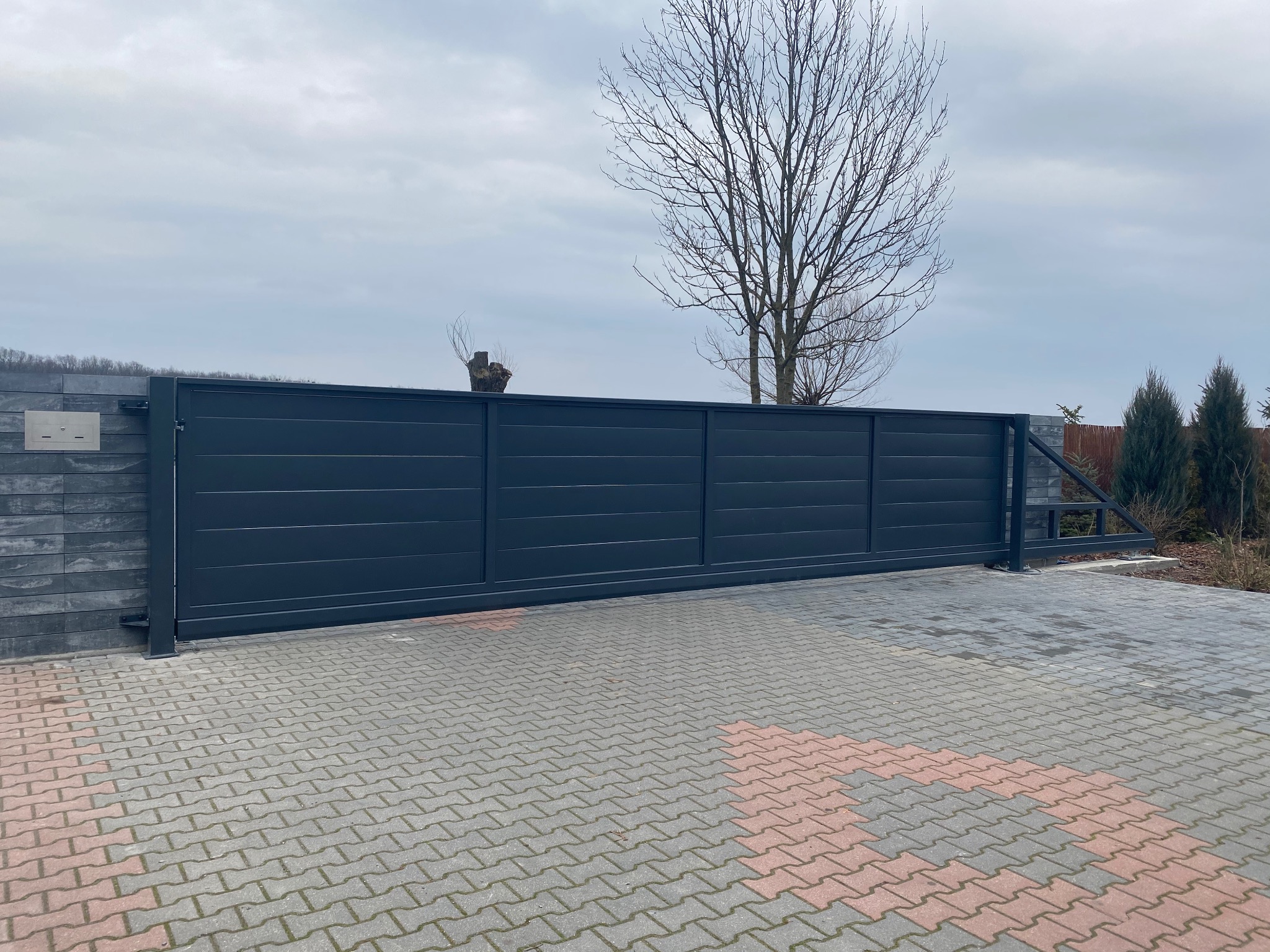 Nowoczesna, przesuwna brama wjazdowa w kolorze grafitowym, z poziomymi panelami, zamontowana na posesji z kostką brukową i minimalistycznym ogrodzeniem z szarej cegły.