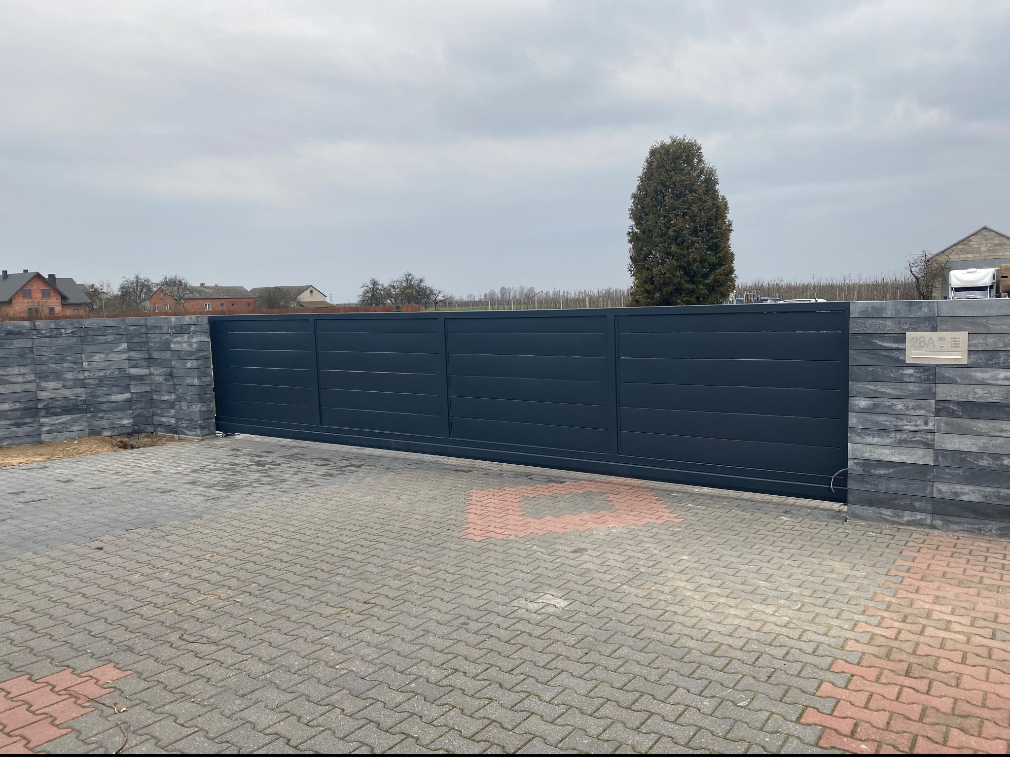Nowoczesna, przesuwna brama wjazdowa w kolorze antracytowym, z poziomymi panelami, zamontowana na posesji z kostką brukową i słupkami z szarej cegły imitującej kamień, widoczna skrzynka na listy...