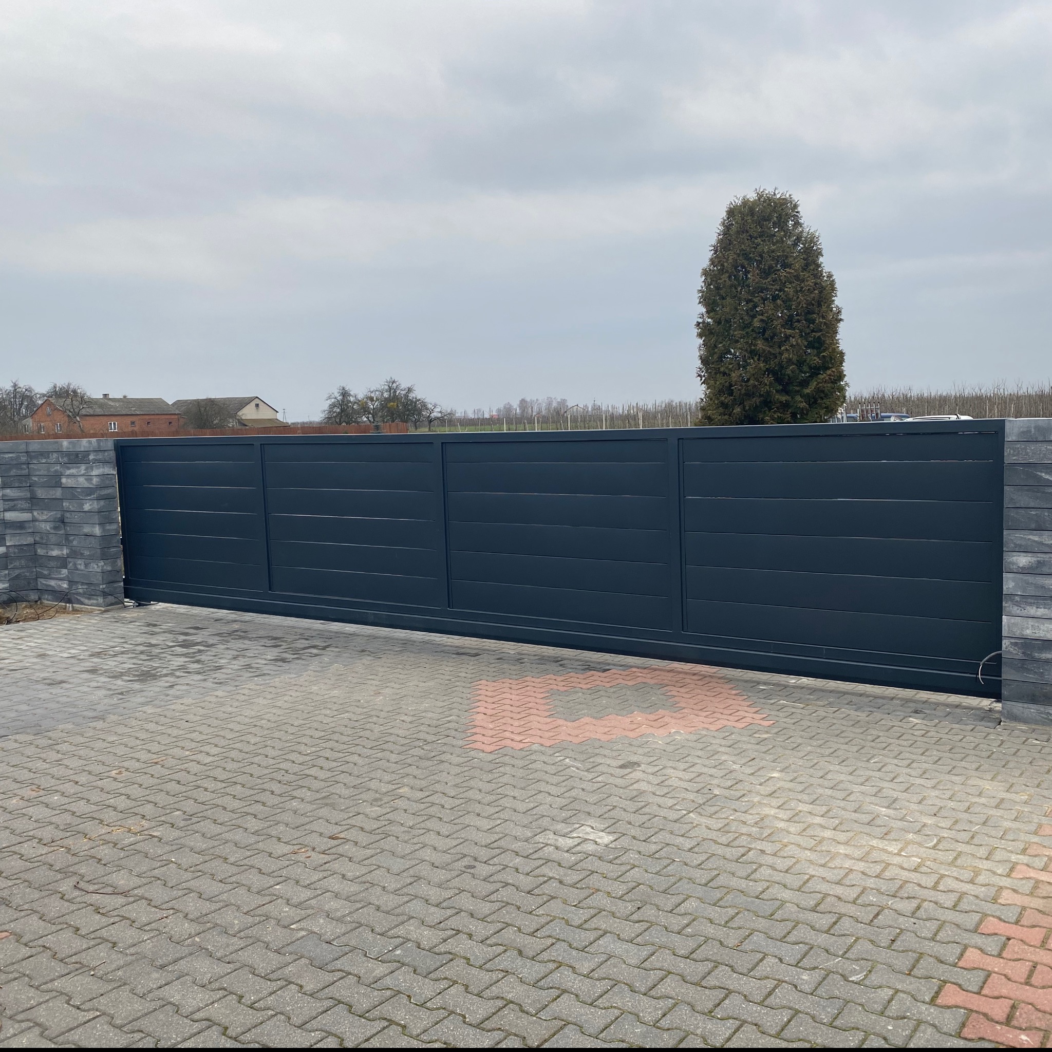 Nowoczesna, przesuwna brama wjazdowa w kolorze grafitowym z poziomymi panelami, zamontowana na posesji z brukowanym podjazdem i widokiem na dom w tle, sfotografowana w pochmurny dzień.