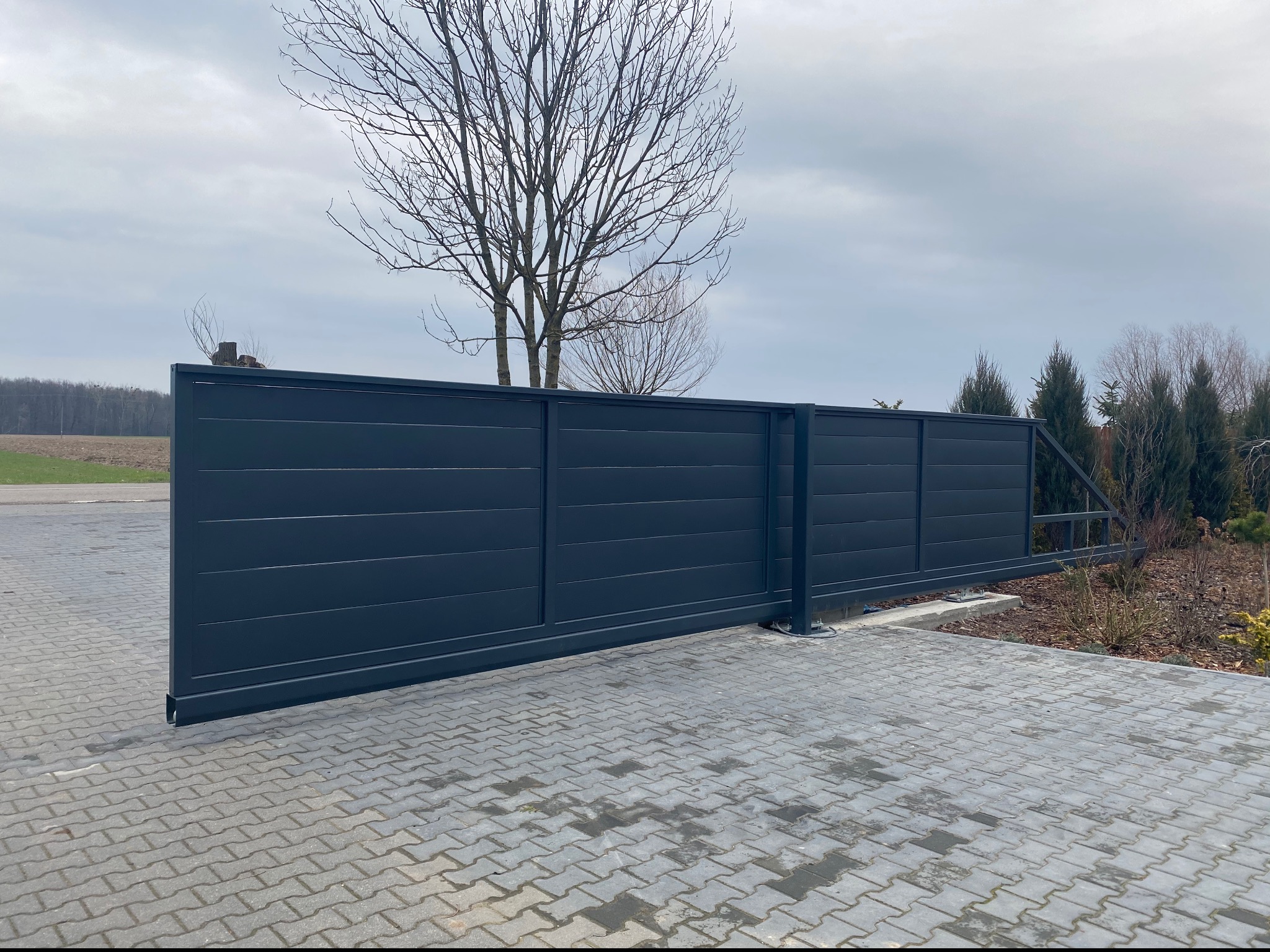 Nowoczesna przesuwna brama wjazdowa w kolorze antracytowym z poziomymi panelami, zamontowana na posesji z kostką brukową i widocznymi drzewami w tle.