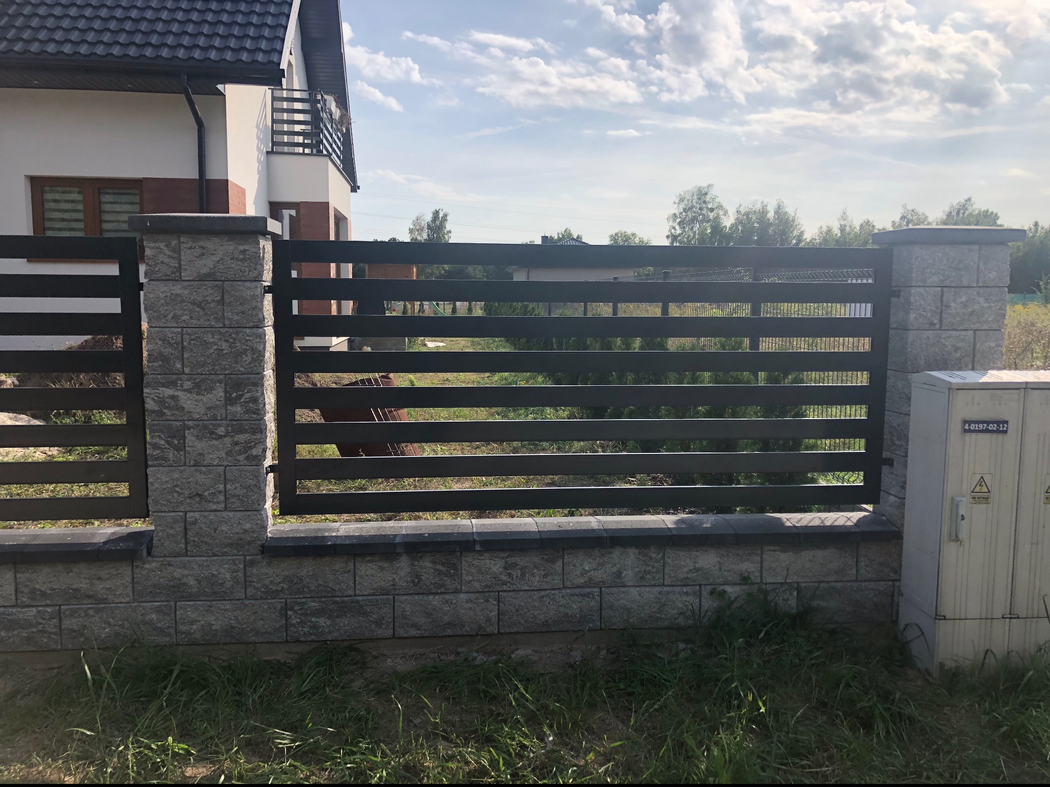 Nowoczesne ogrodzenie z paneli poziomych w kolorze antracytowym, z elementami murowanymi z szarej kostki, na tle domu z balkonem i błękitnego nieba.