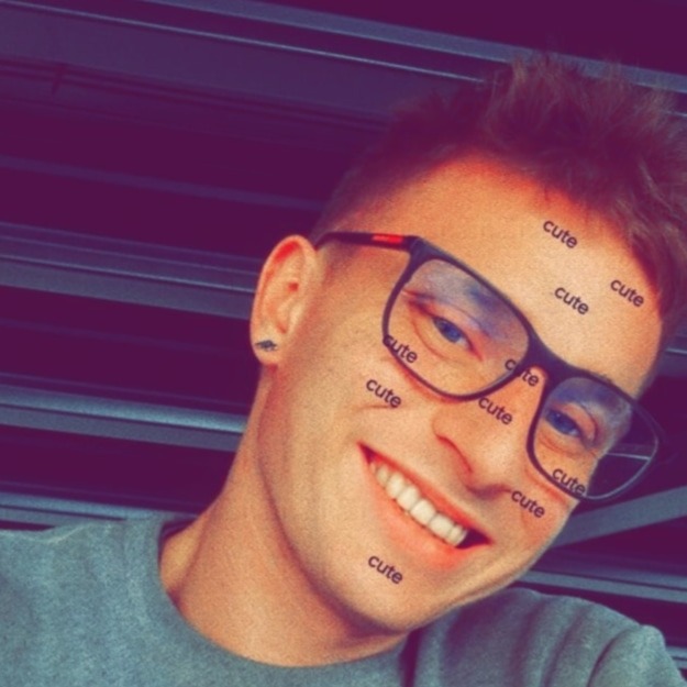 Uśmiechnięty mężczyzna w okularach z filtrem 'cute' na twarzy, szara bluza, widoczne poziome żaluzje w tle.