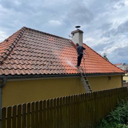 PS&P - Pracownik na drabinie czyści dachówkę z użyciem myjki ciśnieniowej; widoczna różnica w kolorze między umytą a zabrudzoną częścią dachu.