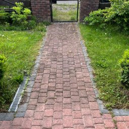 PS&P - Czerwona kostka brukowa prowadząca do metalowej bramki w ceglanym murku, widok na zadbany trawnik i zaparkowany samochód.