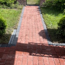 PS&P - Ułożona z czerwonej kostki brukowej ścieżka z szarym obramowaniem prowadząca do metalowej bramki w ceglanym ogrodzeniu, widoczny trawnik i zieleń po bokach, cień na kostce.