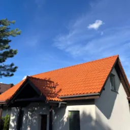 Dom z pomarańczową dachówką ceramiczną na tle błękitnego nieba z pojedynczą chmurą, widoczne rynny i fragment zieleni.