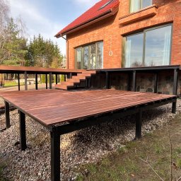 PS&P - Nowoczesny taras kompozytowy z czarną konstrukcją, przylegający do ceglanego domu z dużymi oknami. Widoczne schody i otoczenie z kamieniami.