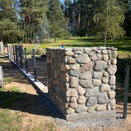 PS&P - Wznoszony mur oporowy z kamienia polnego, z widocznymi metalowymi słupkami i betonową podstawą, na tle zieleni i drzew w słoneczny dzień.