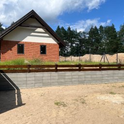 PS&P - Nowoczesny mur oporowy z szarej kostki betonowej i drewnianym płotem na tle domu z czerwonej cegły i zieleni. Widok na pusty plac z piaskiem.