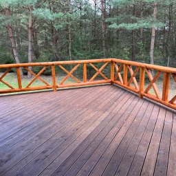 PS&P - Drewniany taras z balustradą w rustykalnym stylu, otoczony lasem. Deski tarasowe ułożone równolegle, balustrada z krzyżowymi elementami. Widok na zielony las w tle.