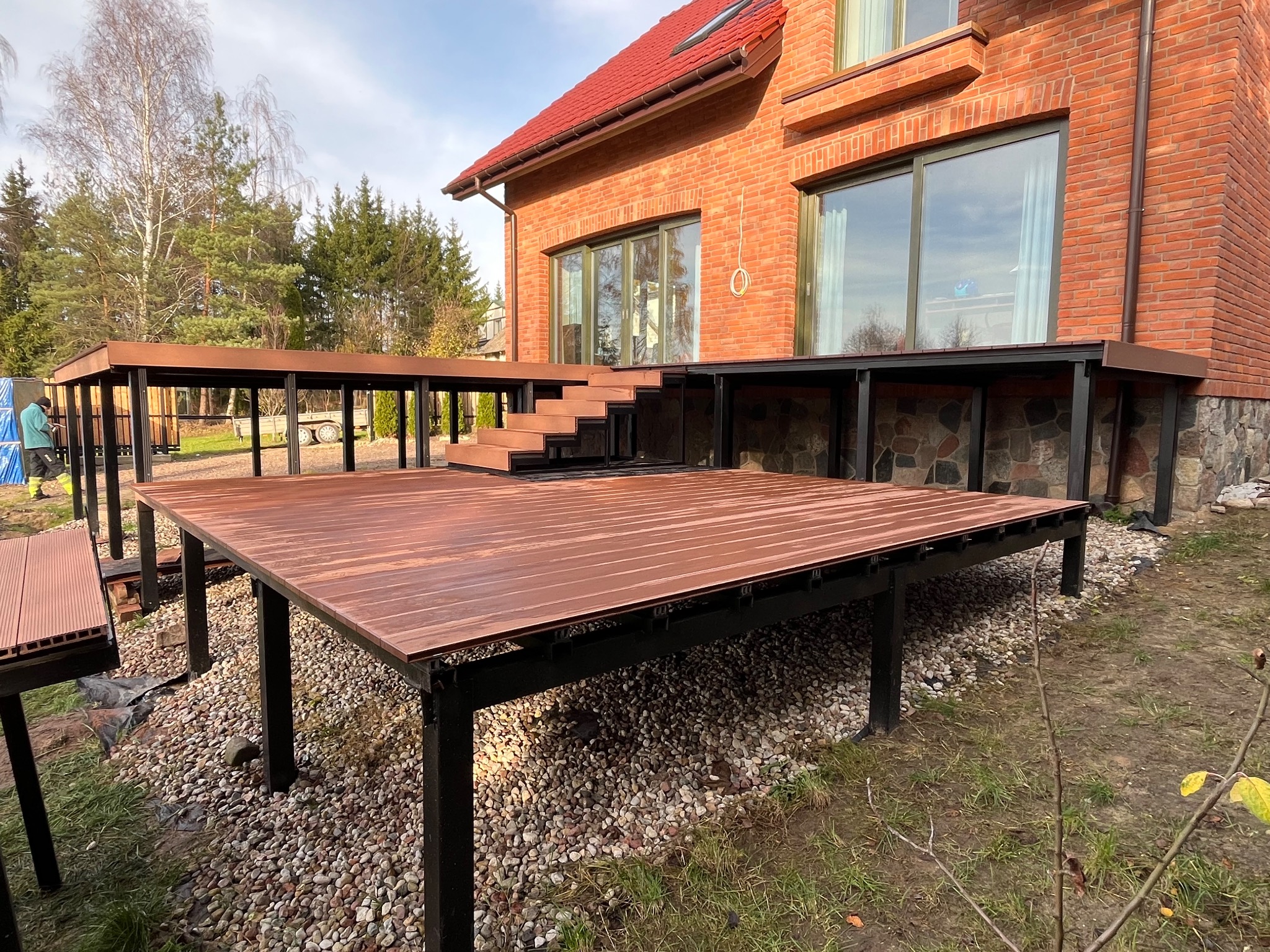 Nowoczesny taras kompozytowy z czarną konstrukcją, przylegający do ceglanego domu z dużymi oknami. Widoczne schody i otoczenie z kamieniami.