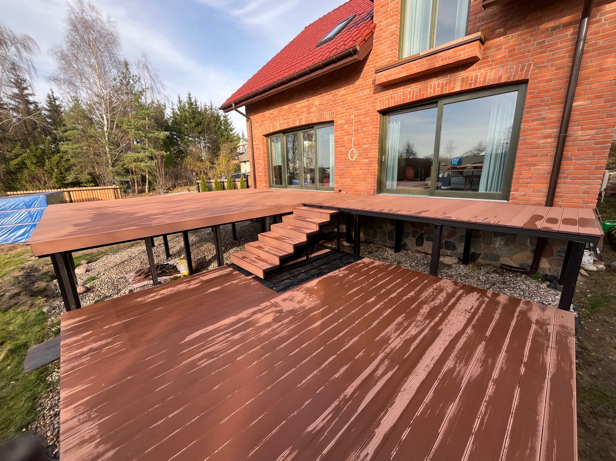 Nowoczesny taras kompozytowy z brązowych desek, schody prowadzące do domu z czerwonej cegły, widok z niskiej perspektywy, częściowo mokra powierzchnia.
