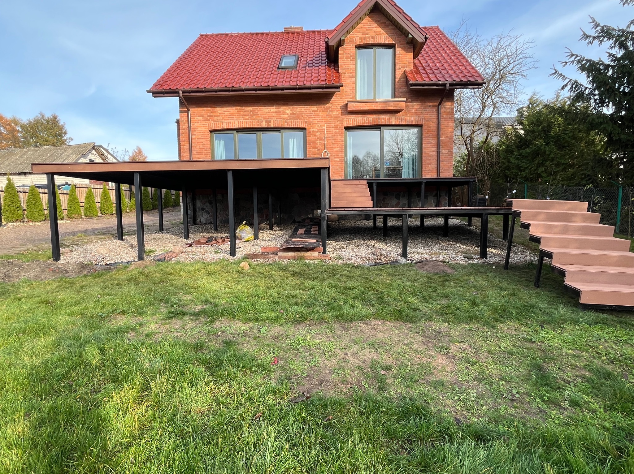 Ceglany dom z czerwonym dachem i tarasem kompozytowym z brązowymi schodami. Widok na trawnik i podjazd z kamienia. Konstrukcja z czarnych słupów.