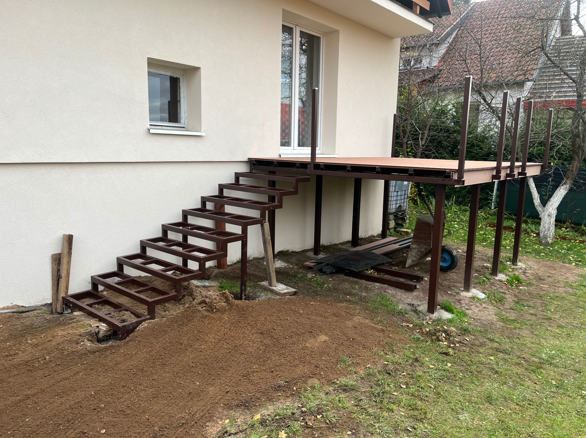 Montaż tarasu kompozytowego z metalową konstrukcją i schodami przy domu. Widoczne elementy konstrukcyjne i przygotowany teren.