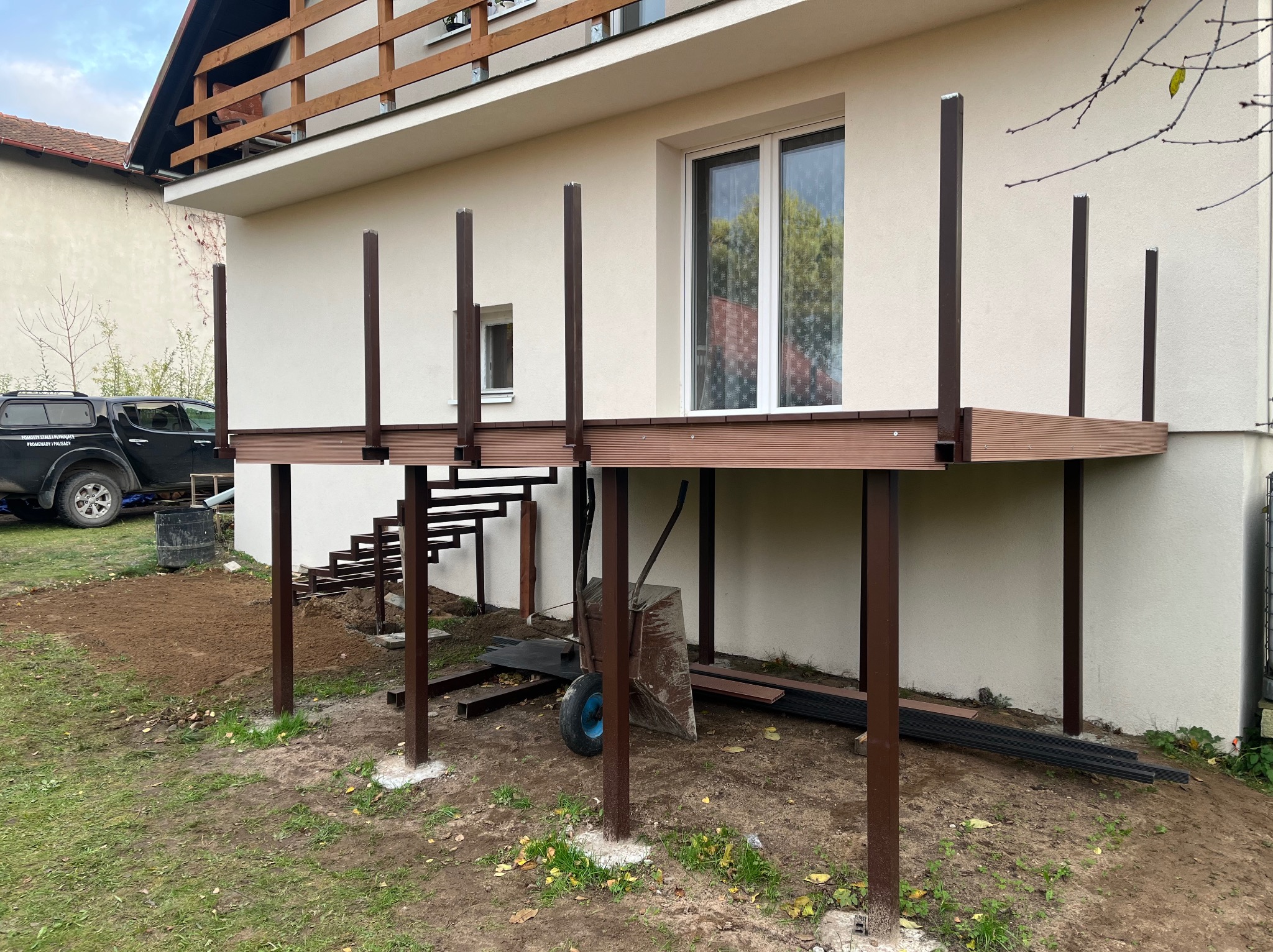 Montaż tarasu kompozytowego: brązowe deski, metalowe słupki, widoczne schody i elementy konstrukcyjne na tle jasnej elewacji domu. Etap budowy, wózek budowlany.