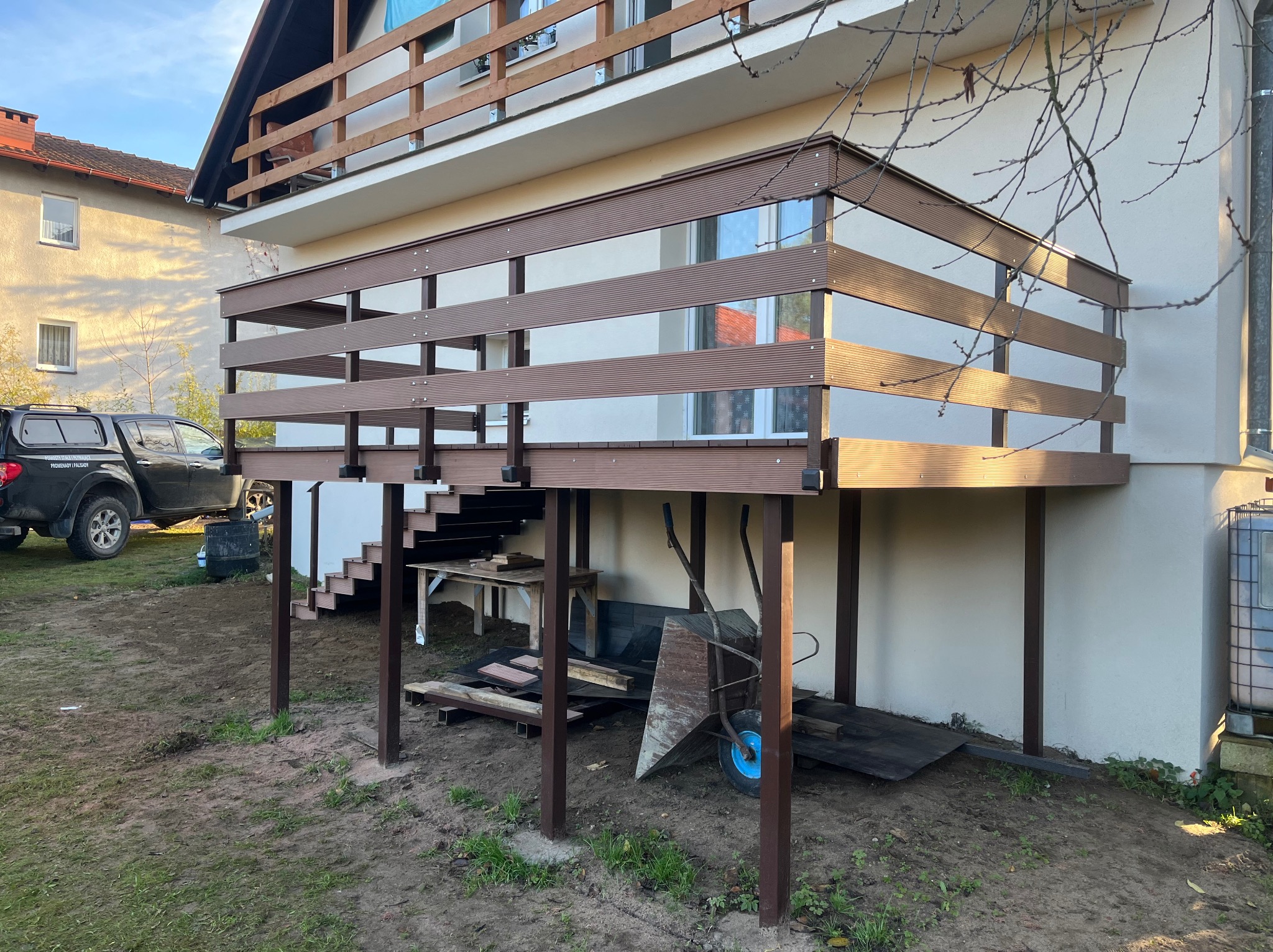 Nowoczesny taras kompozytowy z brązowymi deskami i balustradą, wsparty na słupach. Widoczne schody, teren wokół częściowo zagospodarowany, w tle dom i samochód.