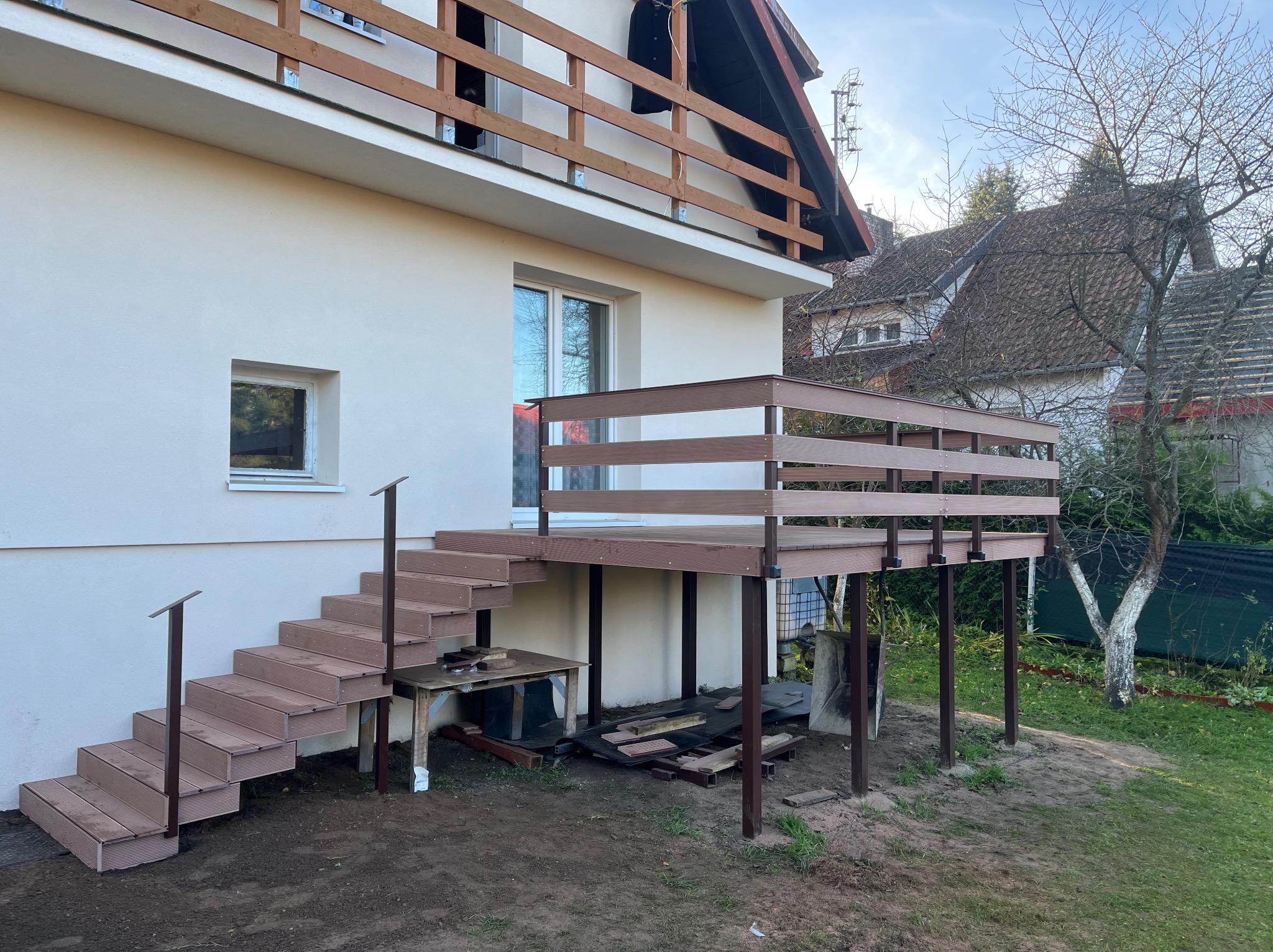 Nowoczesny taras kompozytowy z brązowymi deskami i schodami, przylegający do jasnego budynku. Widoczne podpory i balustrady. Tło stanowi ogród z drzewami.