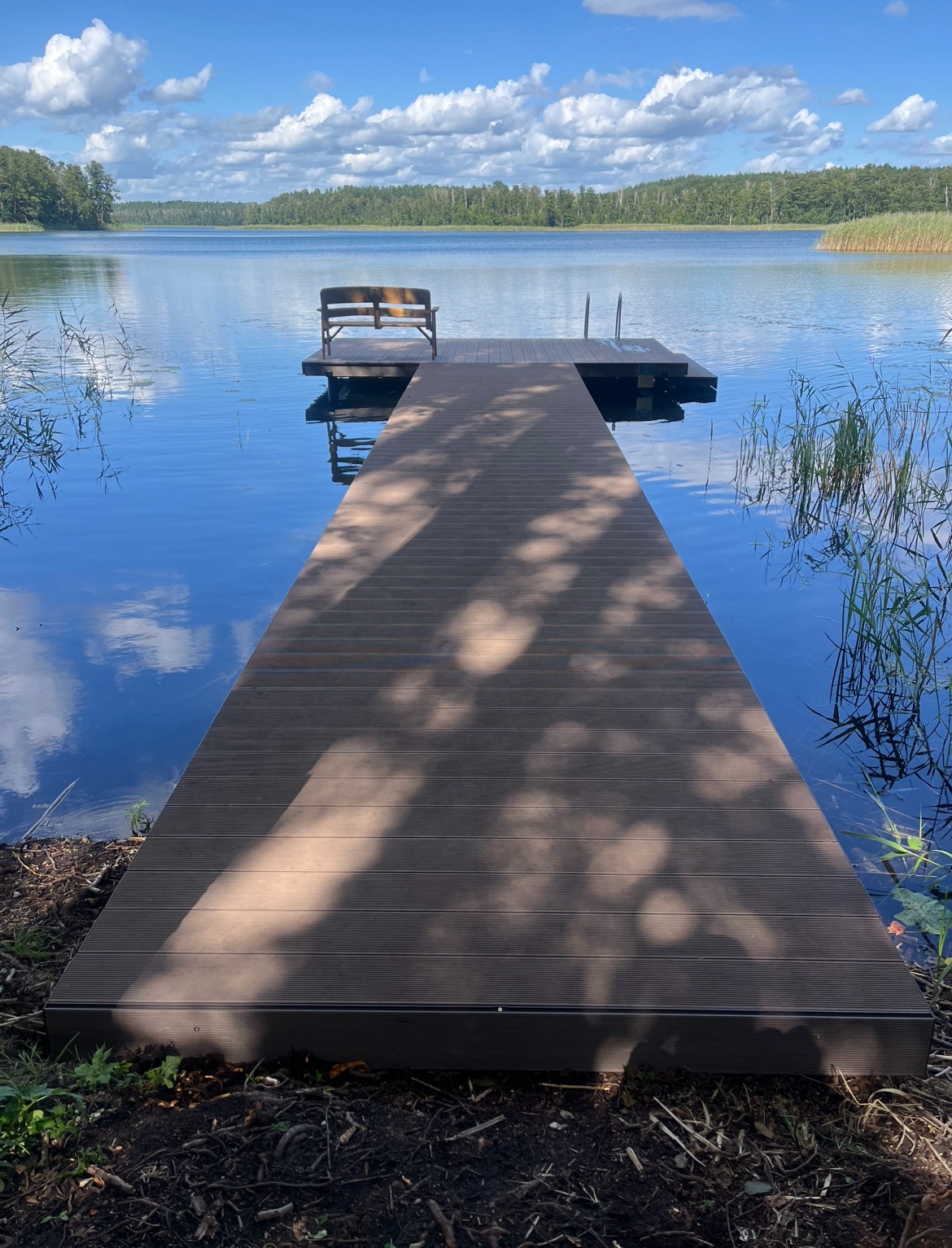 Długi, brązowy pomost z desek kompozytowych prowadzi w głąb spokojnego jeziora. Na końcu pomostu ławeczka i drabinka. Błękitne niebo z chmurami odbija się w wodzie.