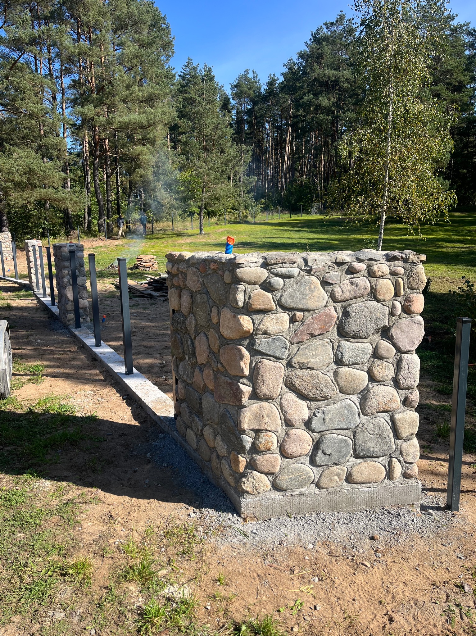 Wznoszony mur oporowy z kamienia polnego, z widocznymi metalowymi słupkami i betonową podstawą, na tle zieleni i drzew w słoneczny dzień.