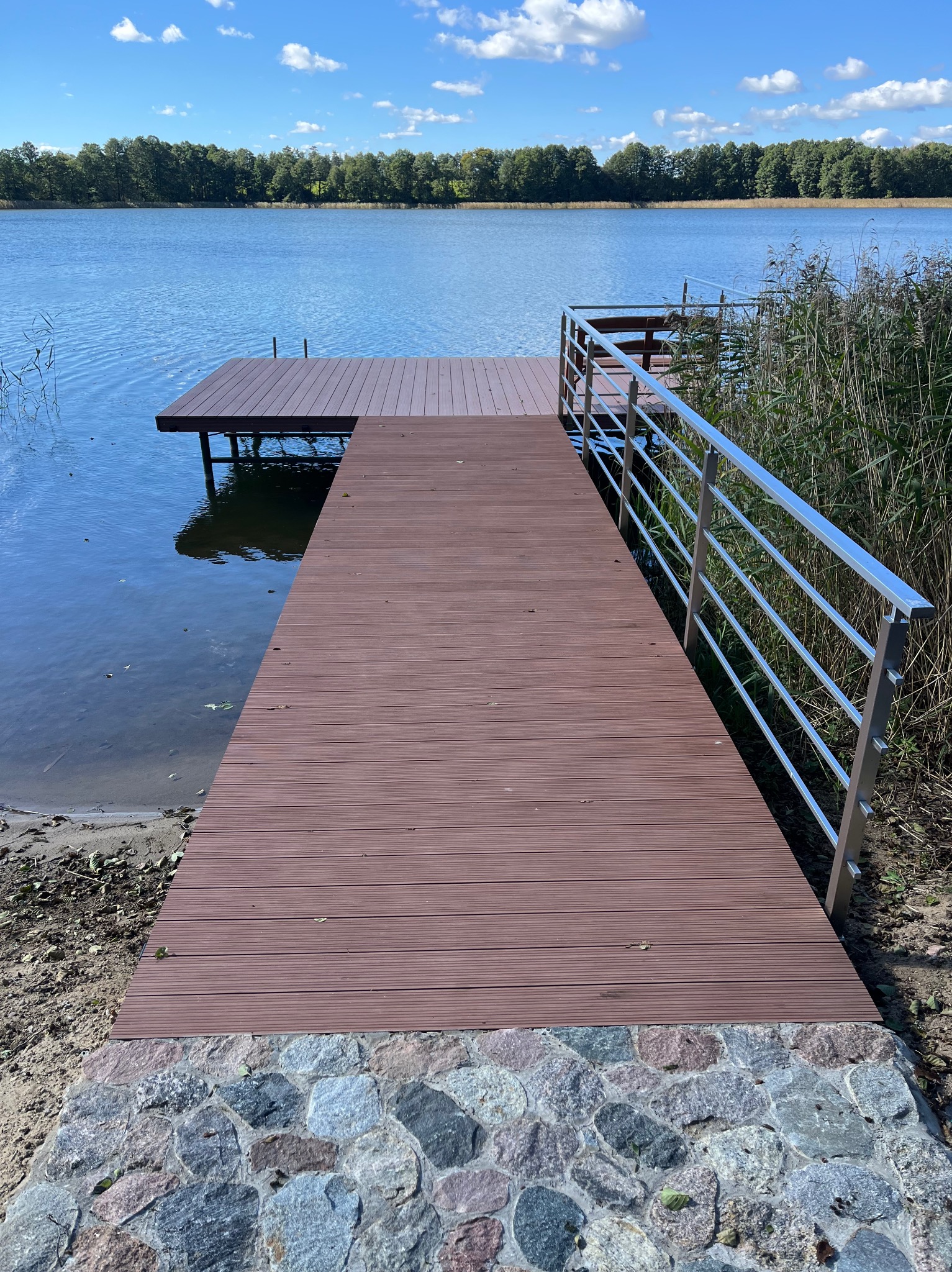 Pomost z desek kompozytowych nad jeziorem, z metalową barierką i kamiennym brzegiem. Widok na wodę i drzewa w tle, słoneczny dzień.