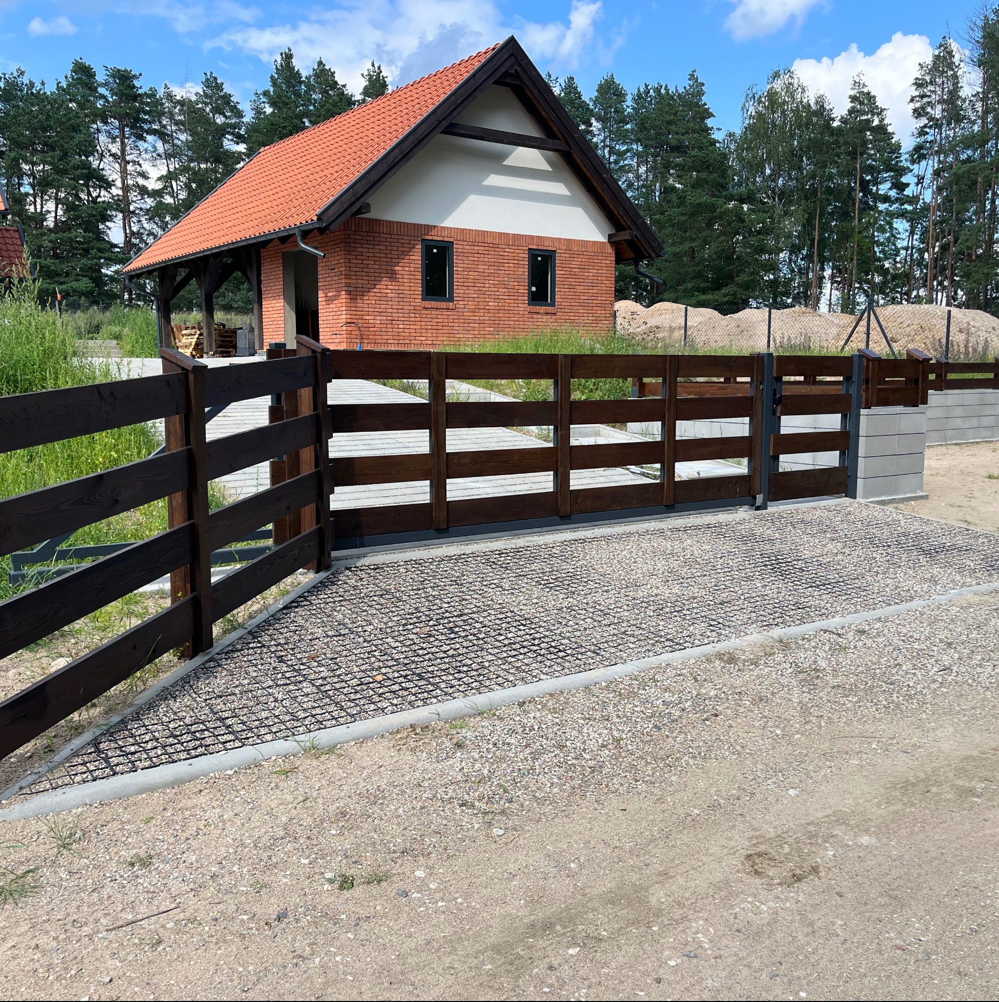 Nowoczesna brama wjazdowa z drewnianymi przęsłami i podjazdem z ażurowych płyt w Pasym. W tle dom z czerwoną dachówką i ceglaną elewacją.