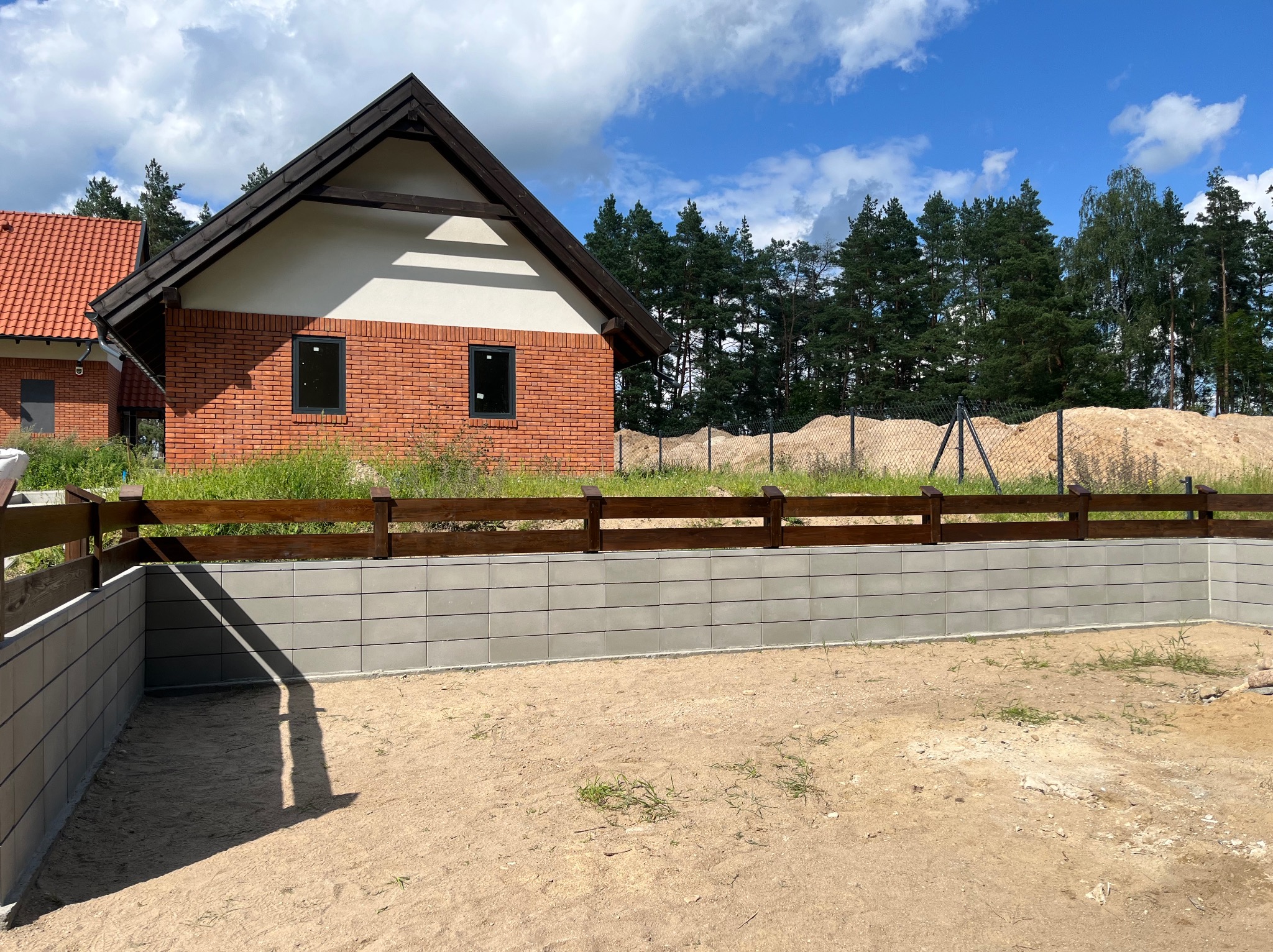 Nowoczesny mur oporowy z szarej kostki betonowej i drewnianym płotem na tle domu z czerwonej cegły i zieleni. Widok na pusty plac z piaskiem.