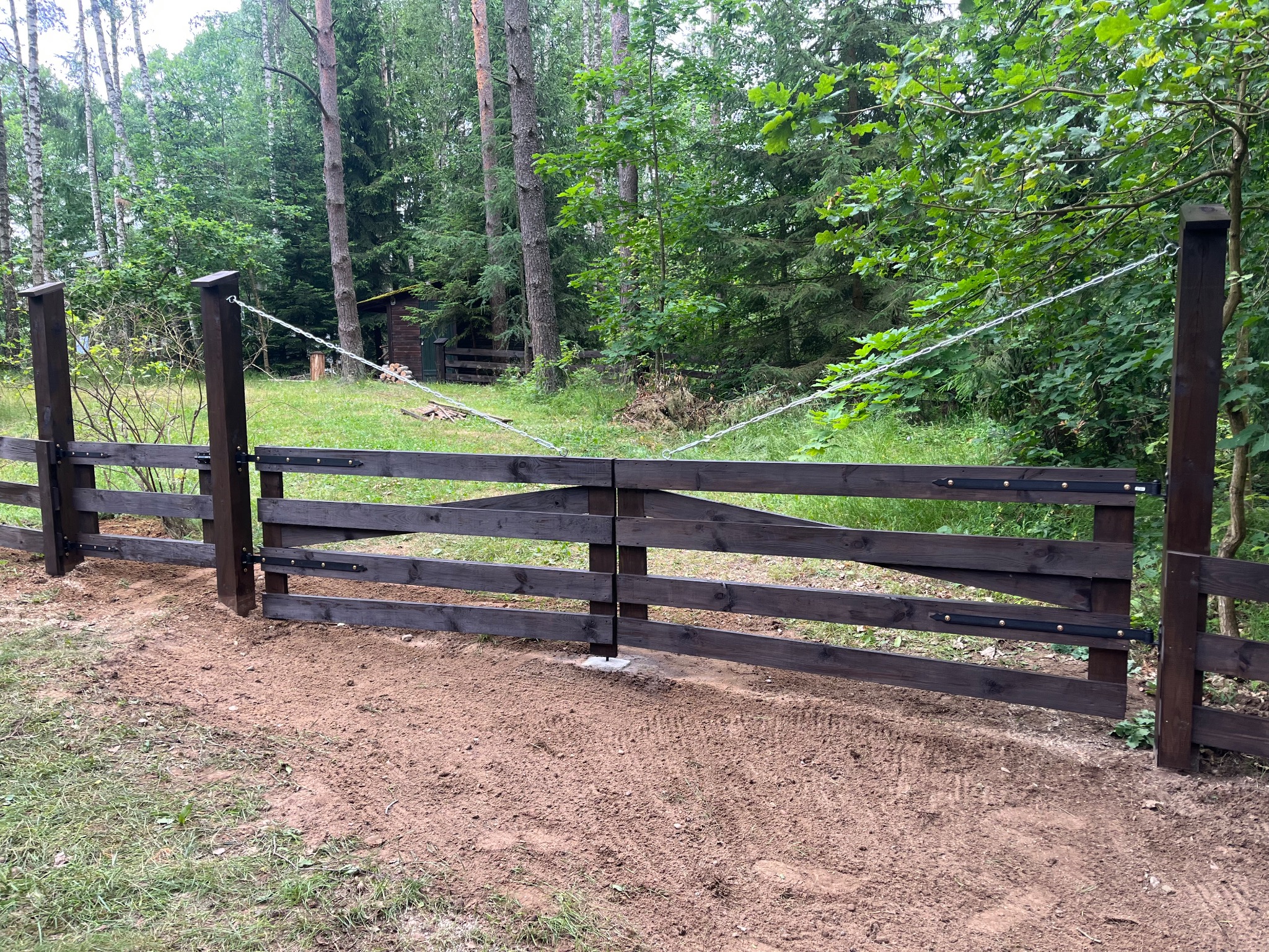 Ciemnobrązowa, drewniana brama wjazdowa z metalowymi okuciami, zabezpieczona łańcuchem, na tle zielonego lasu i trawy. Świeżo wyrównany teren.