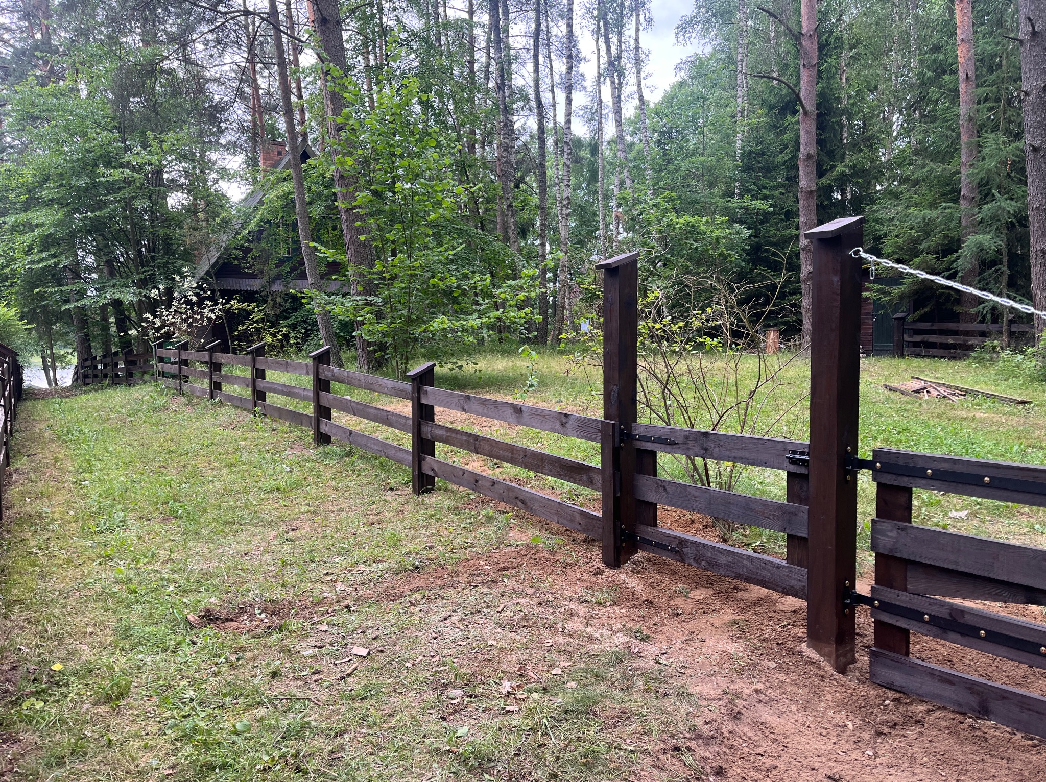 Ciemnobrązowe, drewniane ogrodzenie z furtką na zalesionej działce. Widoczne drzewa i trawa. Furtka otwarta, z łańcuchem. Ujęcie z poziomu gruntu.