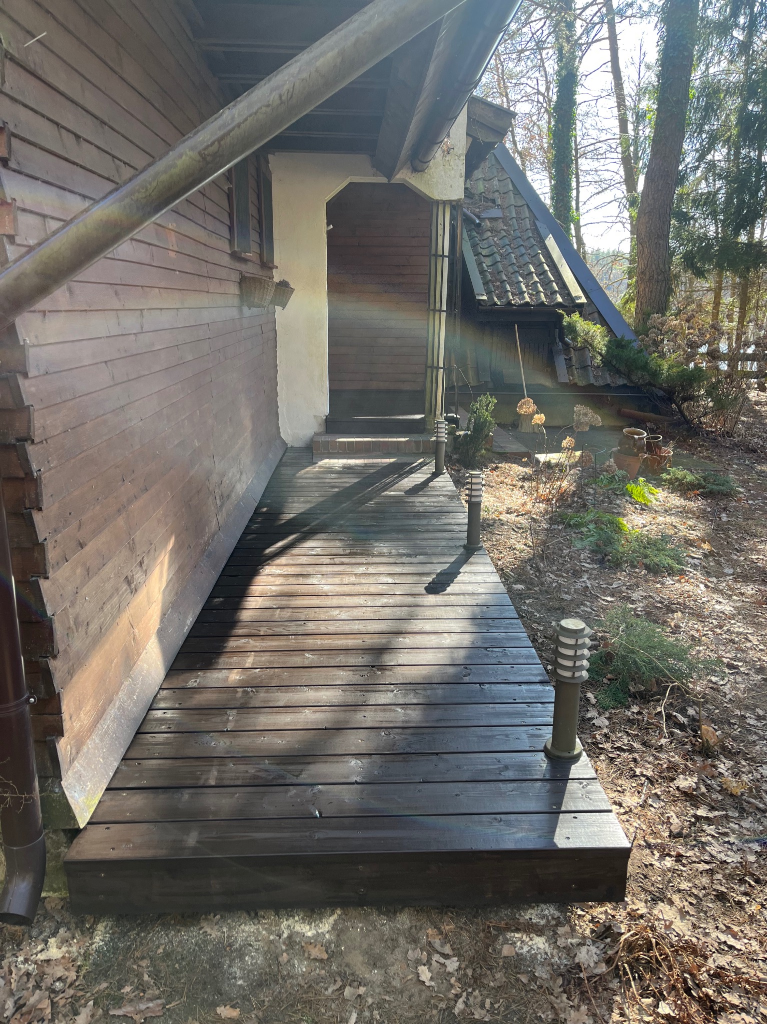 Ciemny, drewniany taras prowadzący do wejścia domu z drewnianą elewacją. Widoczne lampy ogrodowe i fragment dachu. Słoneczny dzień.