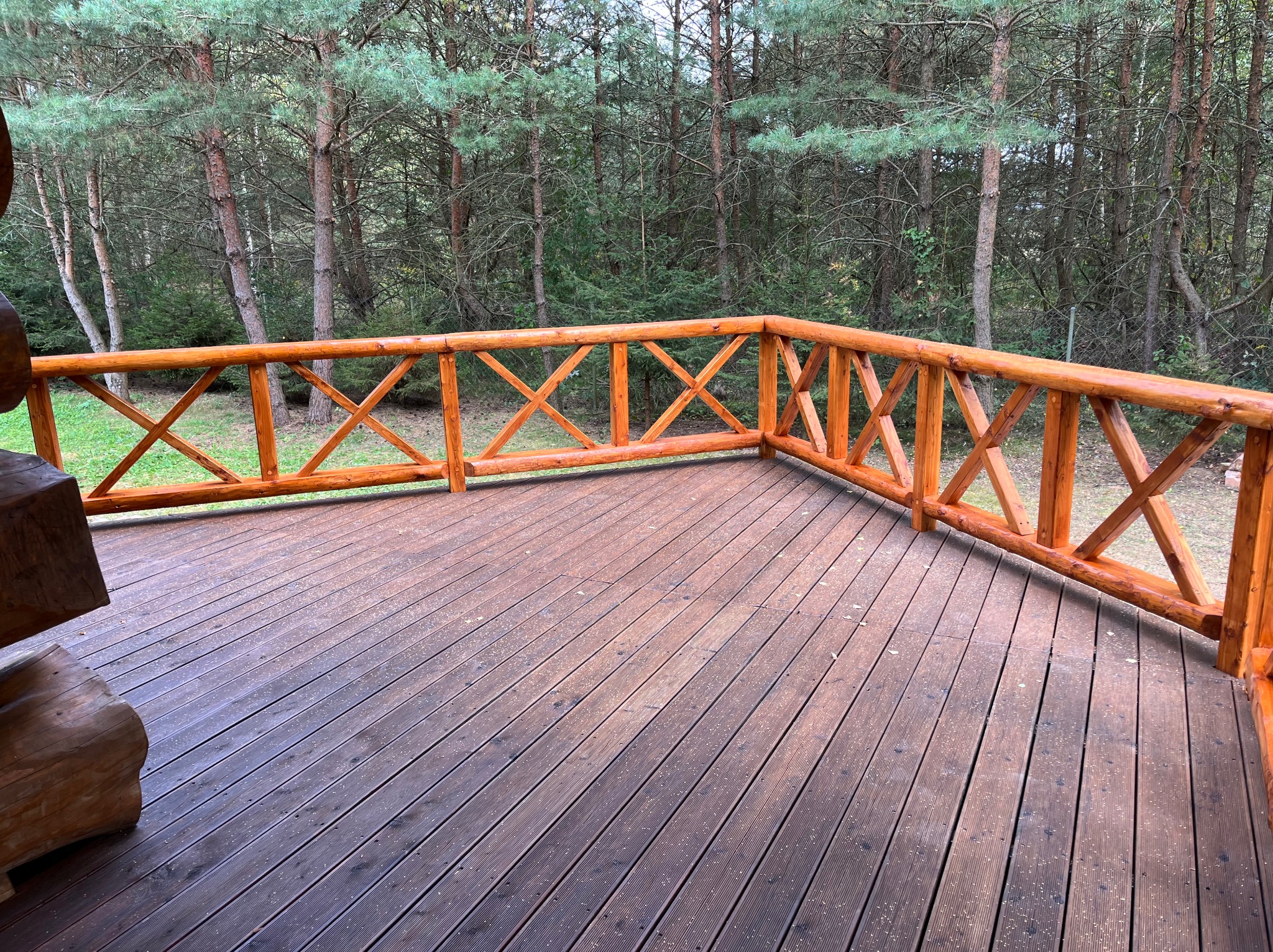 Drewniany taras z balustradą w rustykalnym stylu, otoczony lasem. Deski tarasowe ułożone równolegle, balustrada z krzyżowymi elementami. Widok na zielony las w tle.