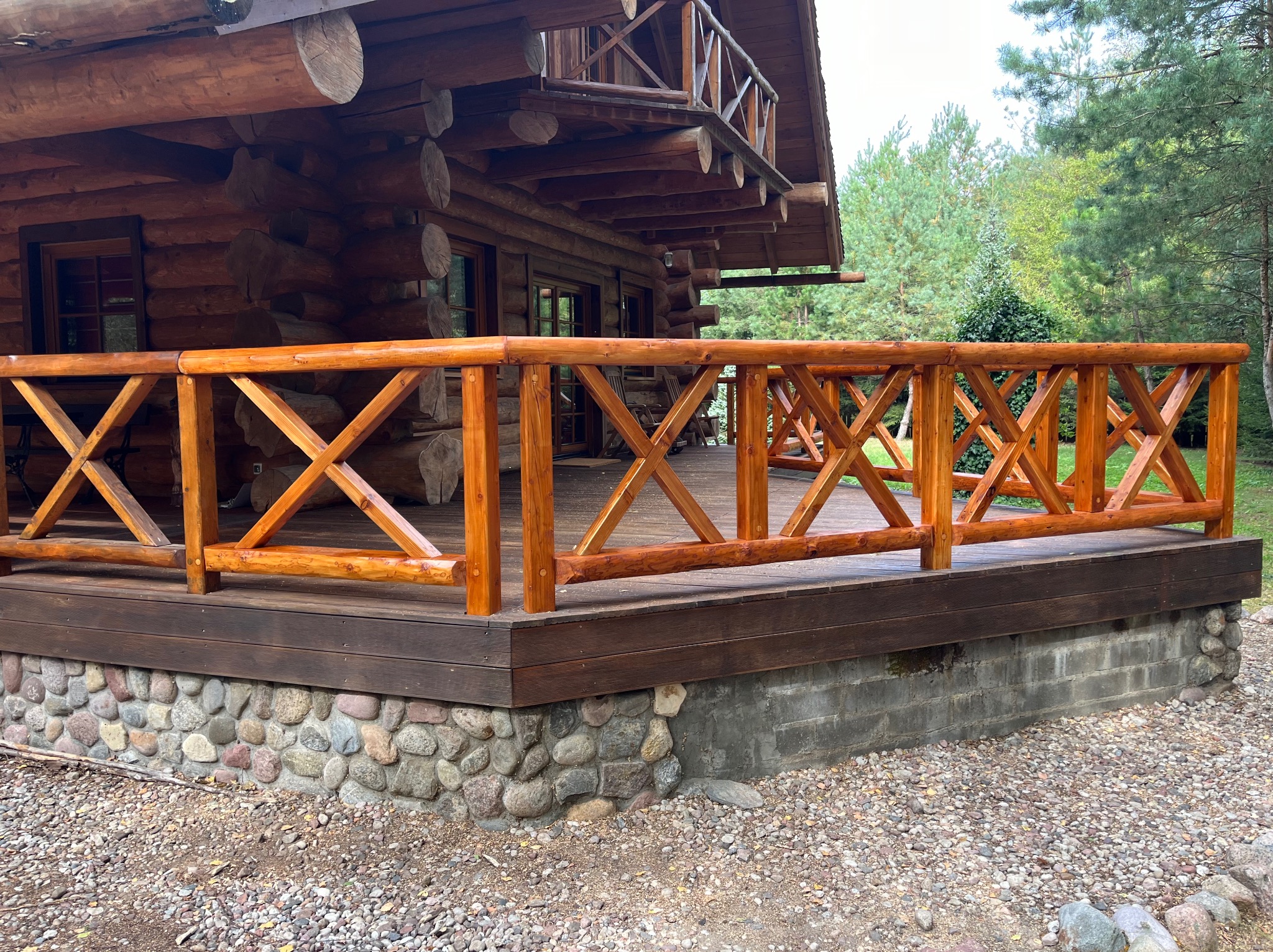 Drewniany taras z balustradą przy domu z bali, podmurówka z kamienia polnego, otoczenie żwirowe, w tle drzewa.