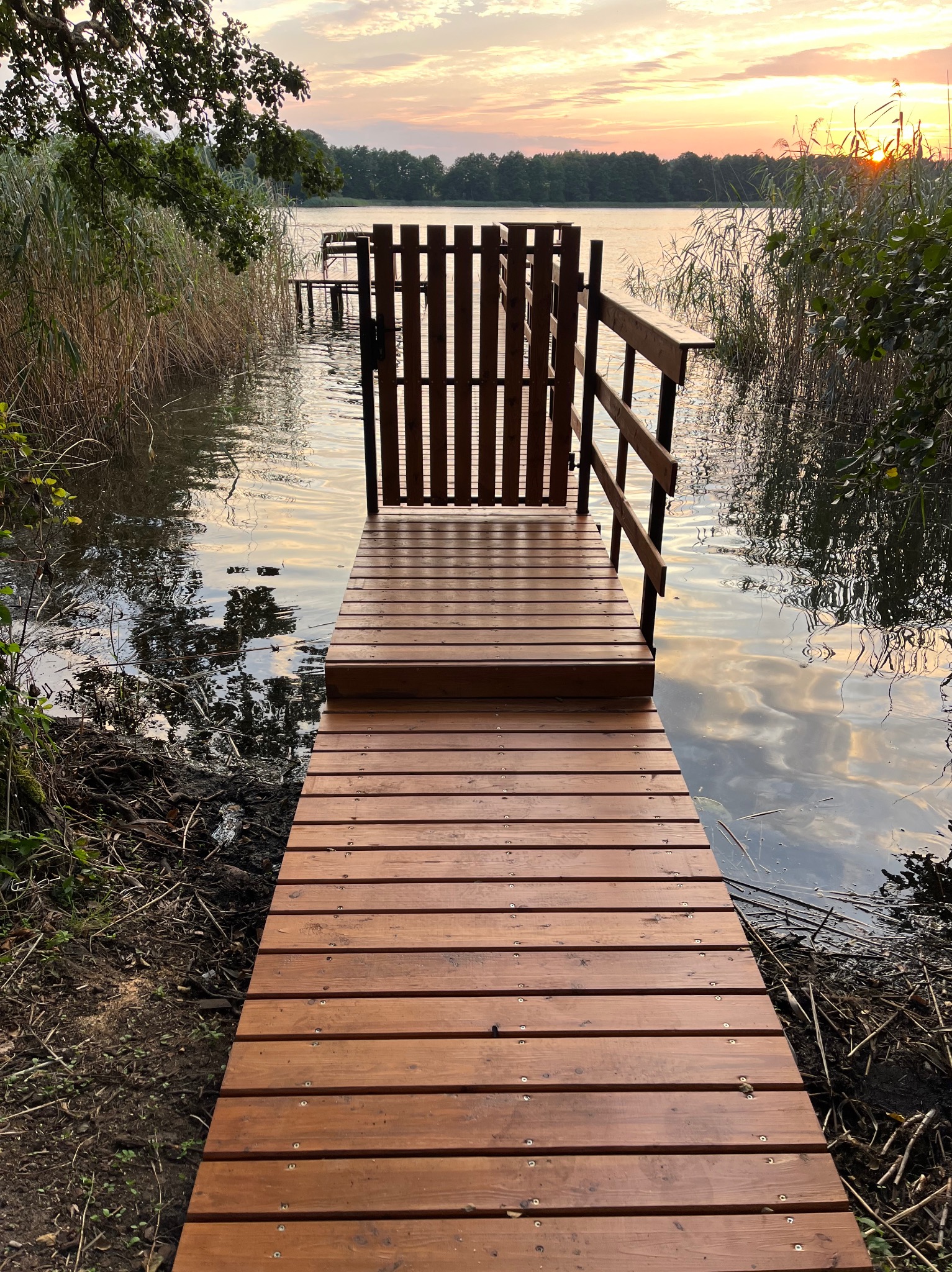 Nowy, drewniany pomost z barierką i furtką, prowadzący do jeziora o zachodzie słońca. Deski pomostu w ciepłym odcieniu brązu, w tle trzciny i spokojna tafla wody.