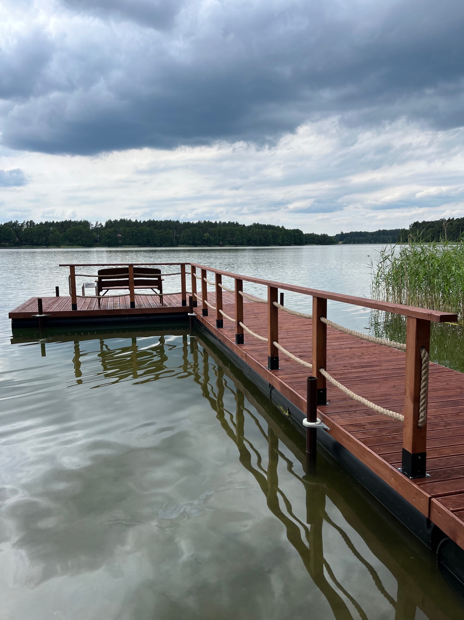 Drewniany taras nad jeziorem z ławką i barierkami z linami. Widok na spokojną wodę i pochmurne niebo. Nowoczesny design tarasu.