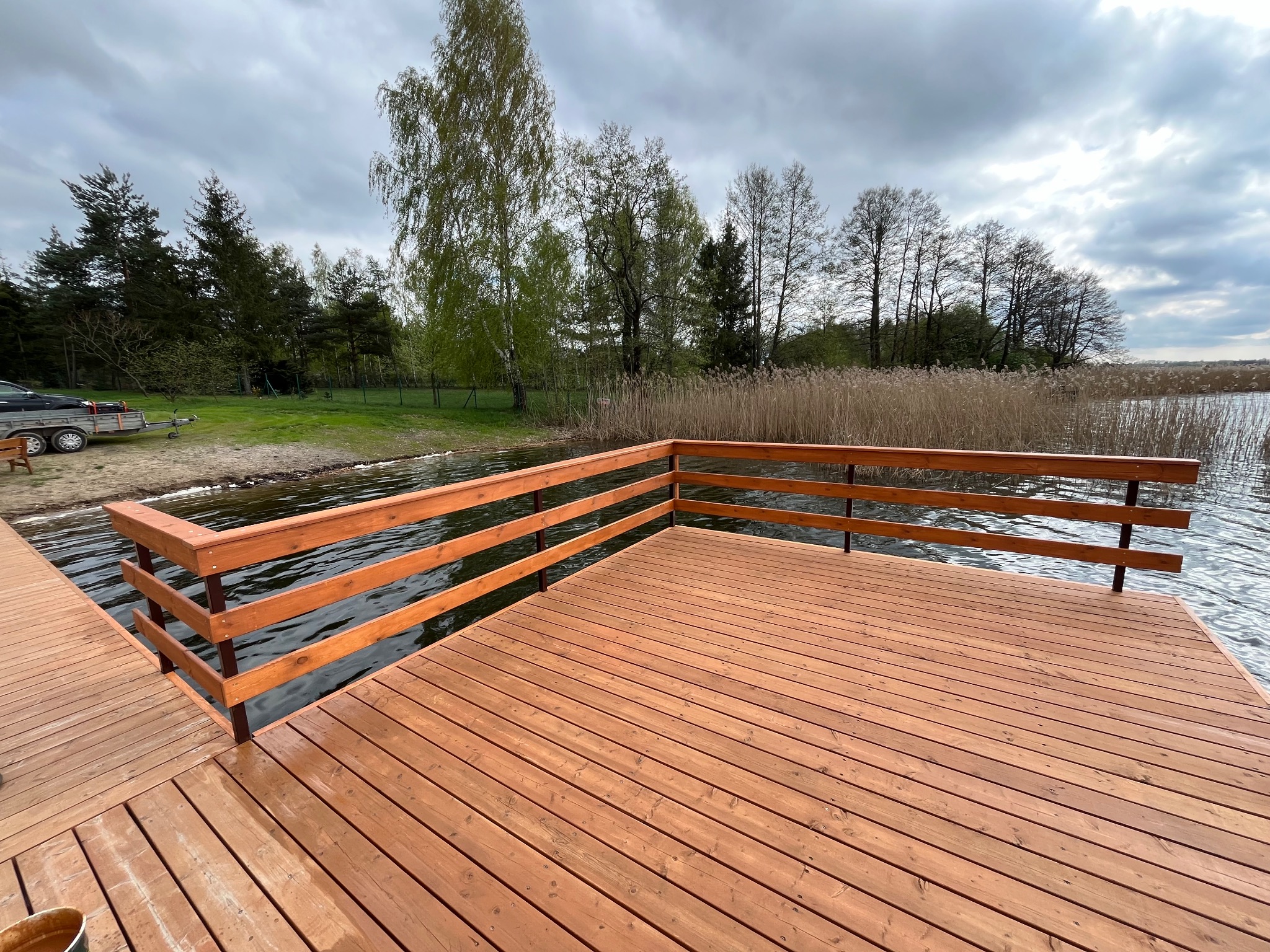 Nowy, drewniany taras nad jeziorem z brązową balustradą. Widoczne deski tarasowe i woda. W tle drzewa i pochmurne niebo. Świeżo zaimpregnowane drewno.