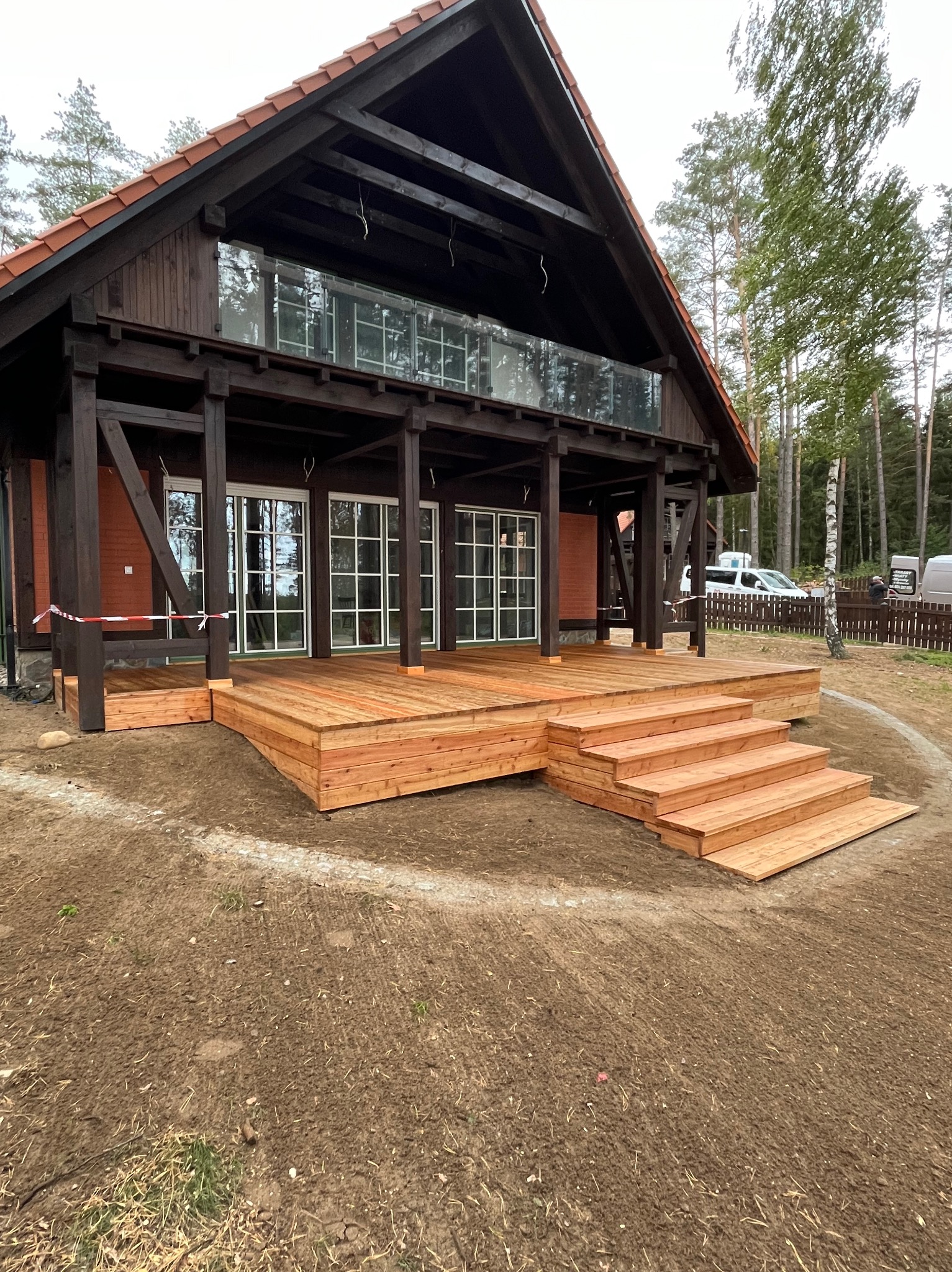Nowy, drewniany taras z jasnego drewna, z niskimi schodami, przylegający do ciemnego, drewnianego domu z przeszkloną balustradą. Widok z poziomu gruntu.