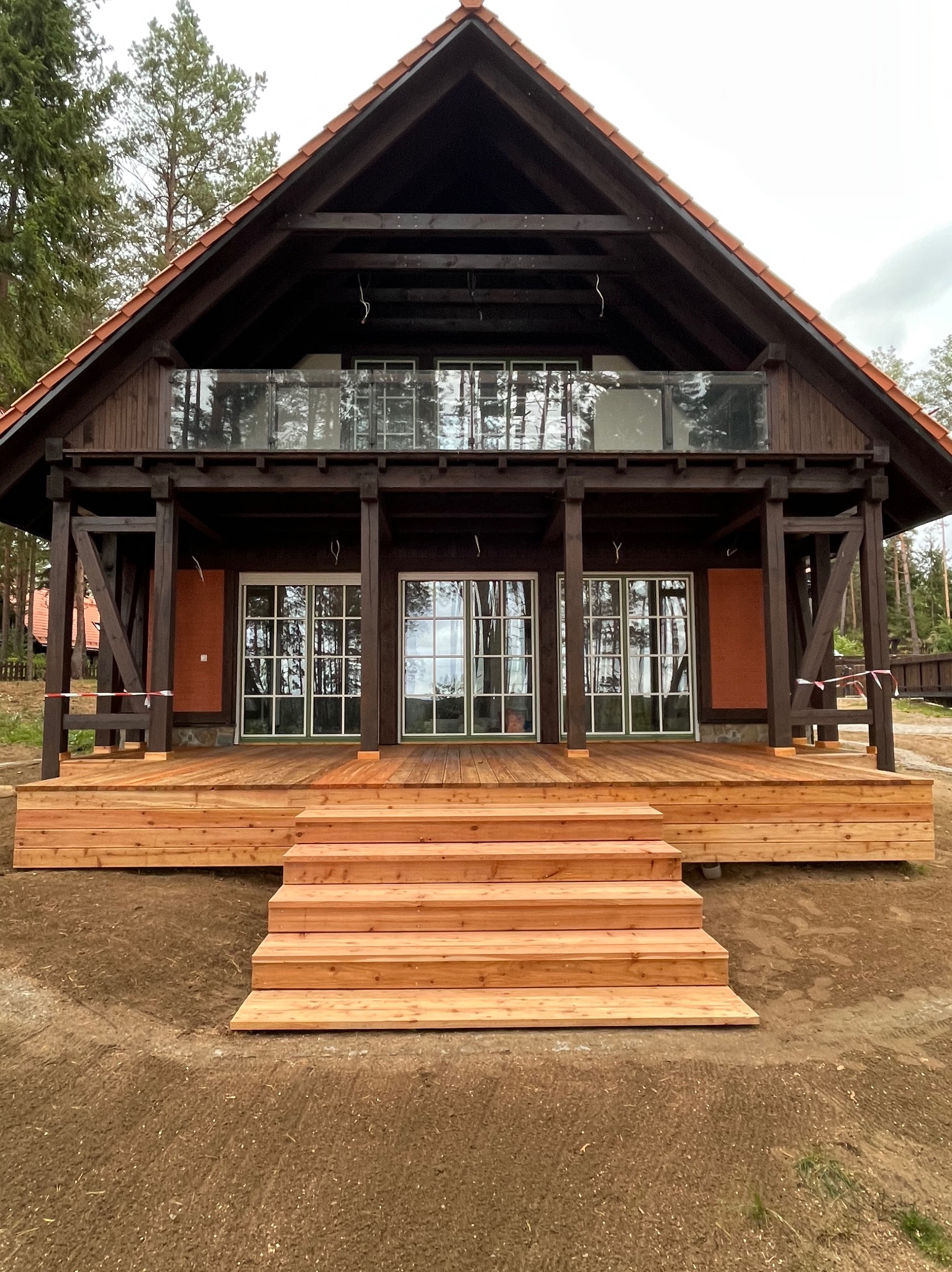 Nowy, jasny taras drewniany z schodami przed domem z ciemnego drewna. Widoczny balkon z szklaną balustradą. Świeżo usypana ziemia wokół tarasu.