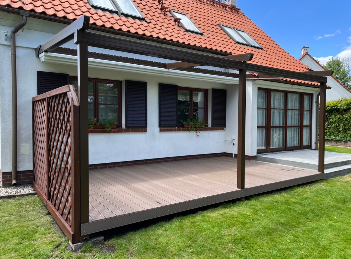 Zadaszony taras kompozytowy z brązową konstrukcją i ażurowym panelem bocznym, przylegający do białego domu z brązowymi okiennicami i czerwonym dachem.